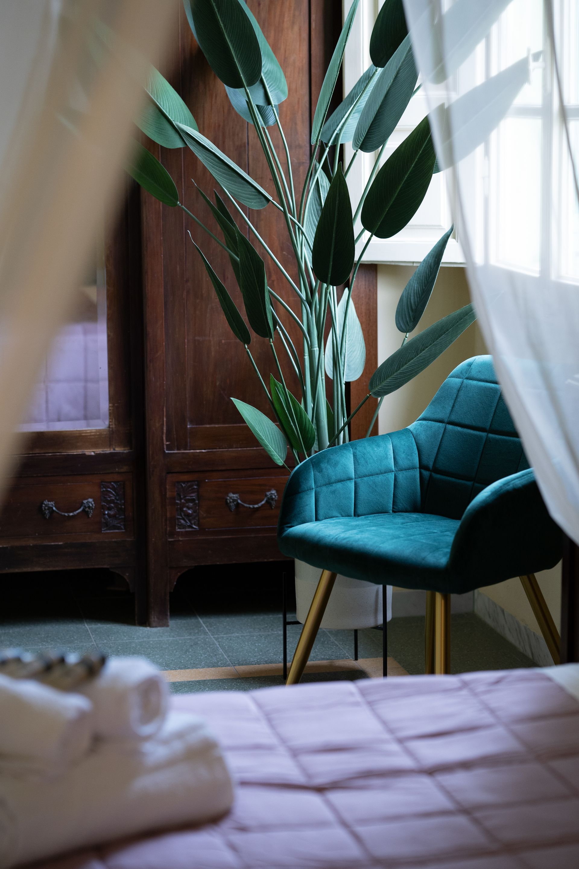 Teal armchair near a tall green plant and dark wooden cabinet; soft focus on bed.