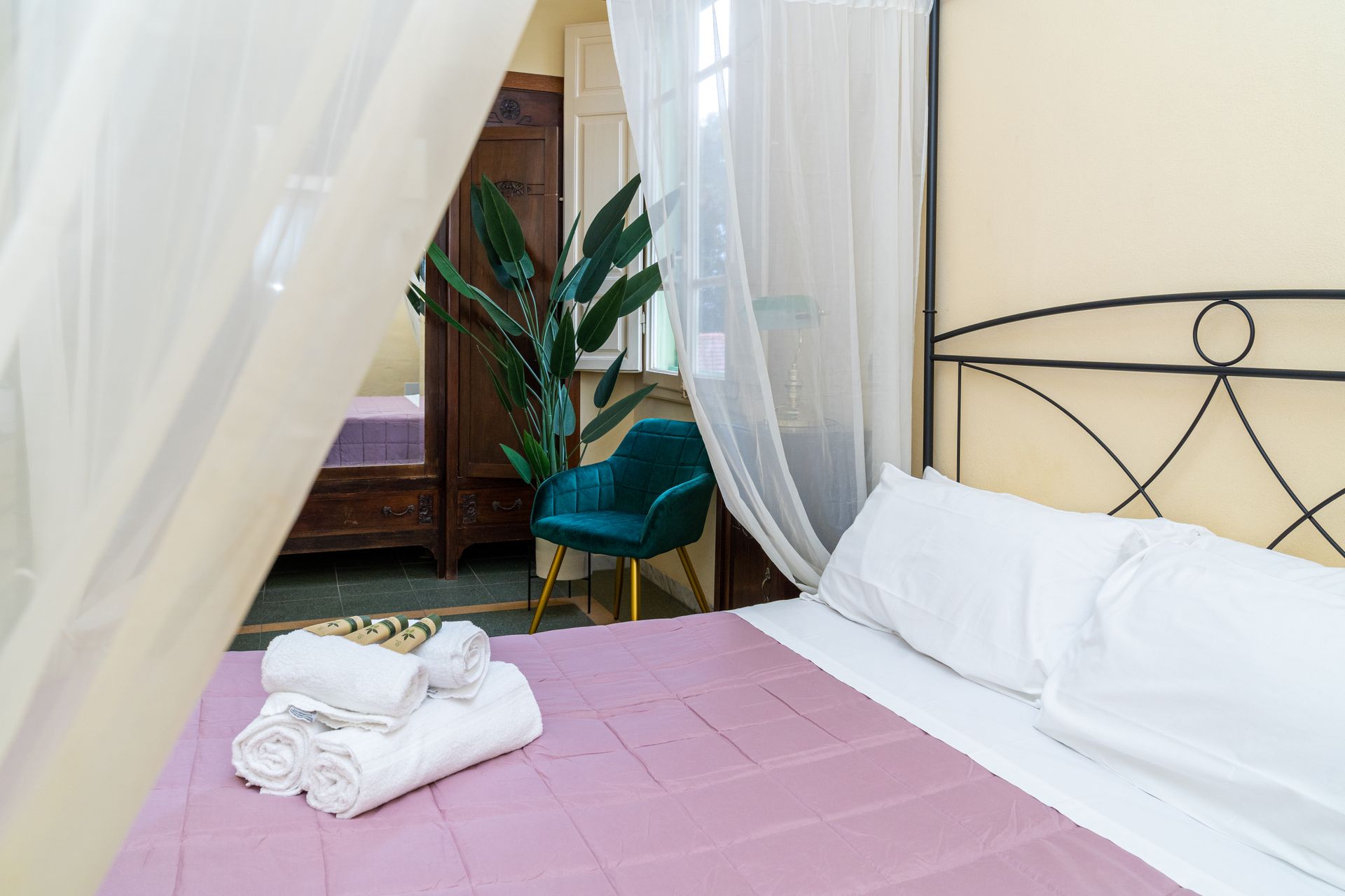 Bedroom with bed, white linens, lavender bedspread, and towels. Green chair, wooden cabinet, and sheer curtains.