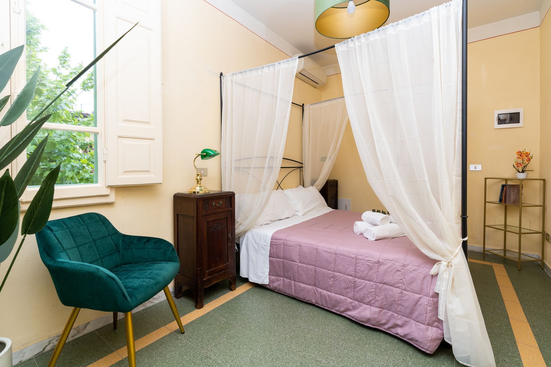 Bedroom with four-poster bed, teal chair, and small side table. Light-colored walls and green flooring.