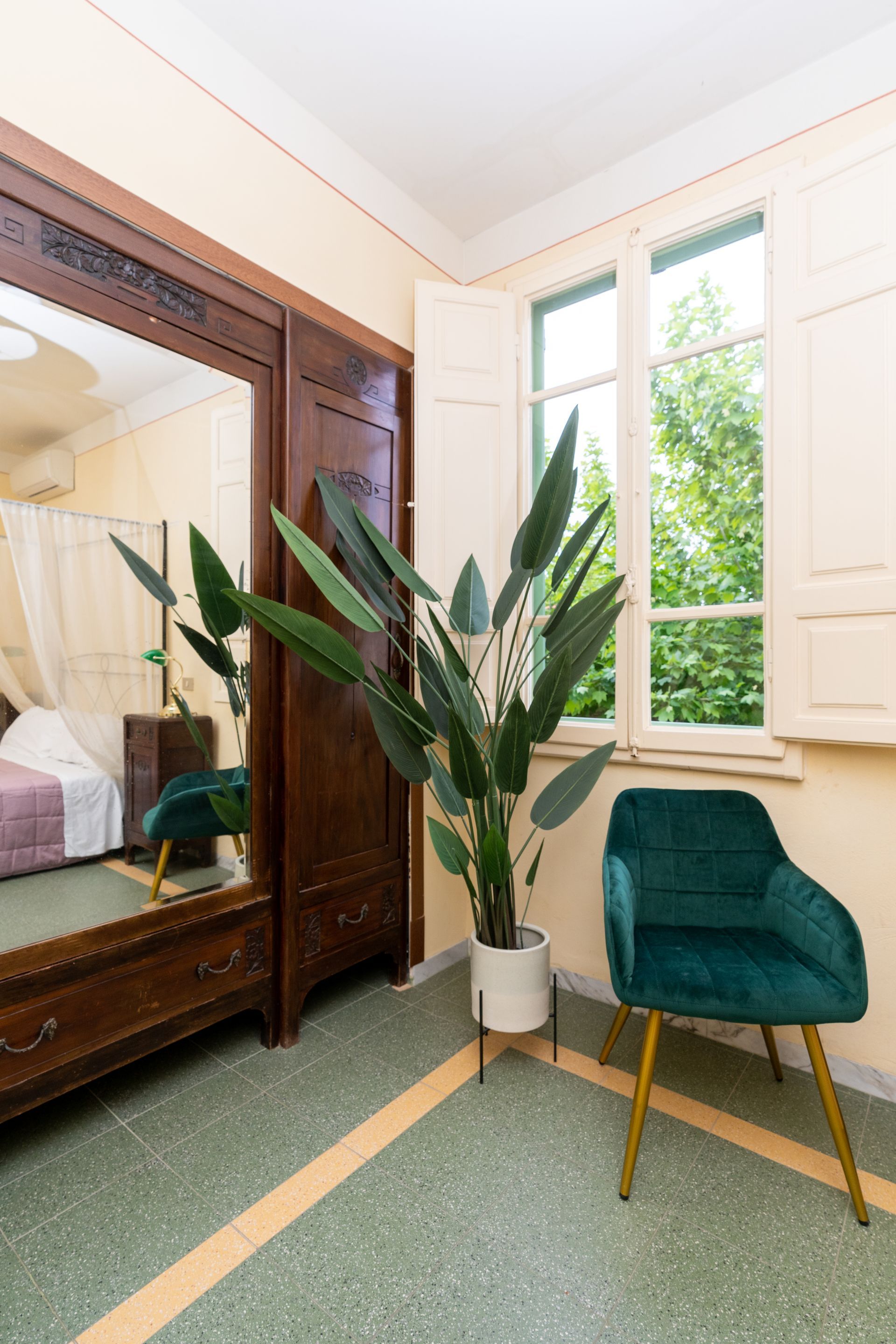 Bedroom corner with a tall plant, teal chair, window with shutters, and a large mirror.