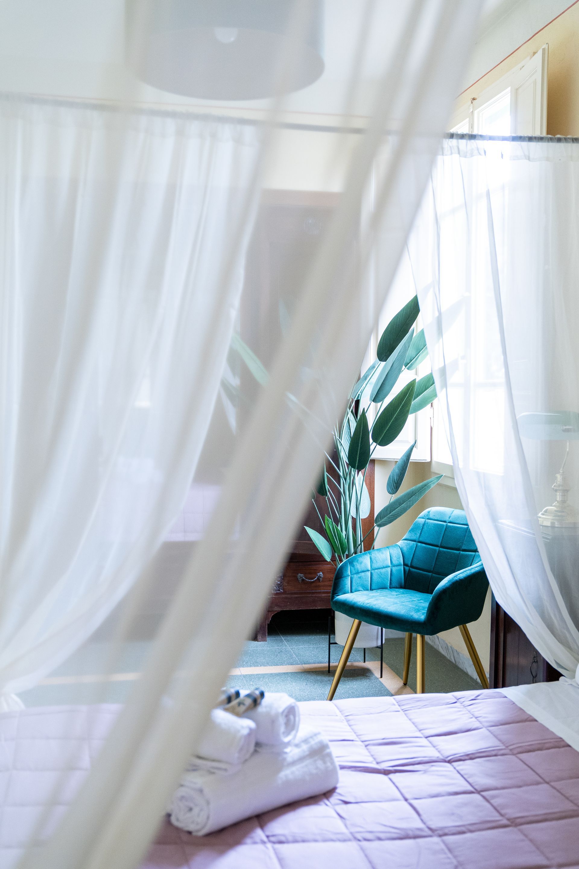 A bed draped with sheer curtains in a room with a teal chair and a plant by a window.