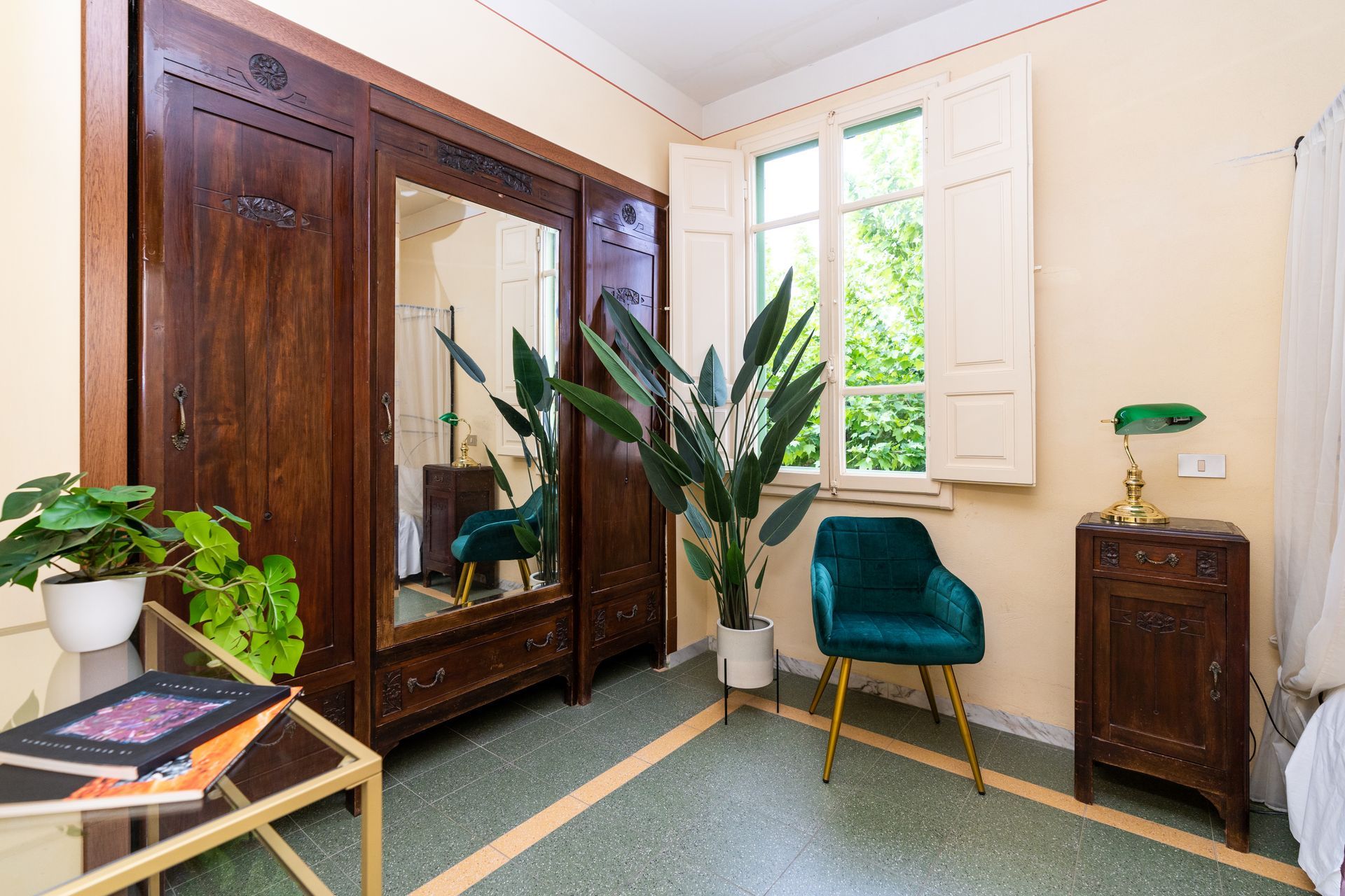 Bedroom with dark wood wardrobe, emerald chair, and plant. Window with shutters.
