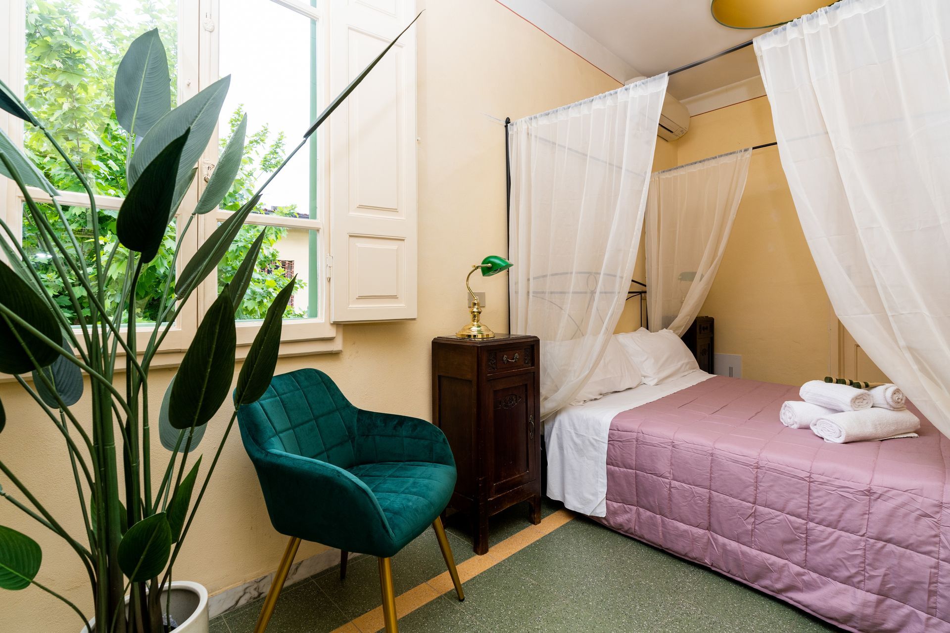Bedroom with bed, canopy, green velvet chair, and large potted plant.