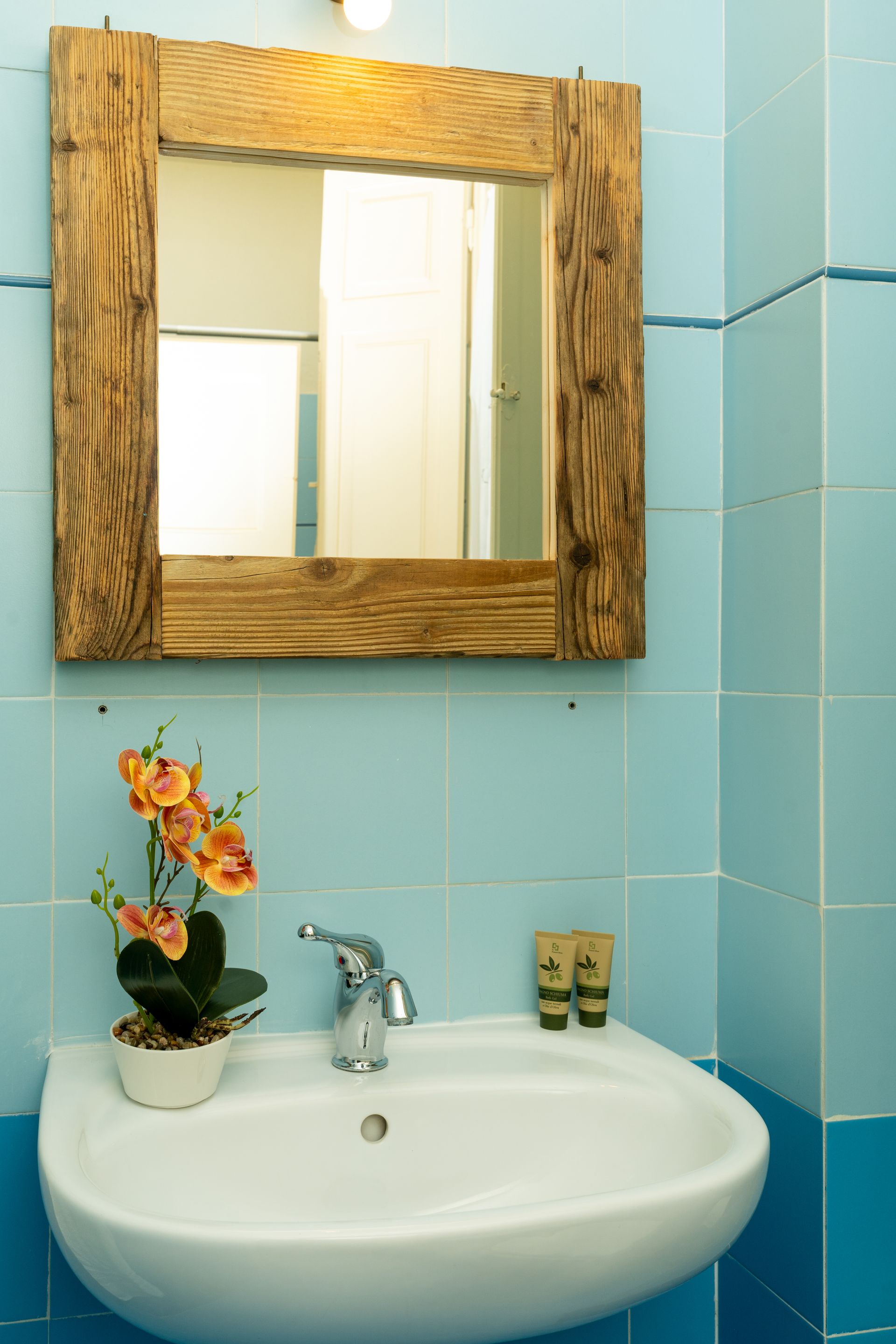 Bathroom sink with mirror in a light blue-tiled room. Artificial flowers sit on the counter.