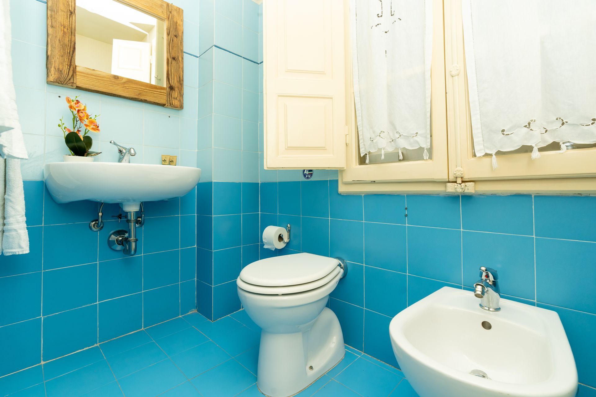 Blue-tiled bathroom with a toilet, bidet, sink, mirror, and a window with curtains.