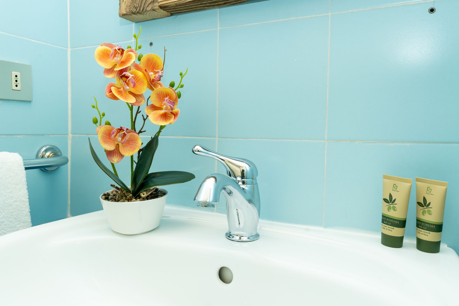 White sink with chrome faucet, orange flower, and two green tubes against blue tiled wall.