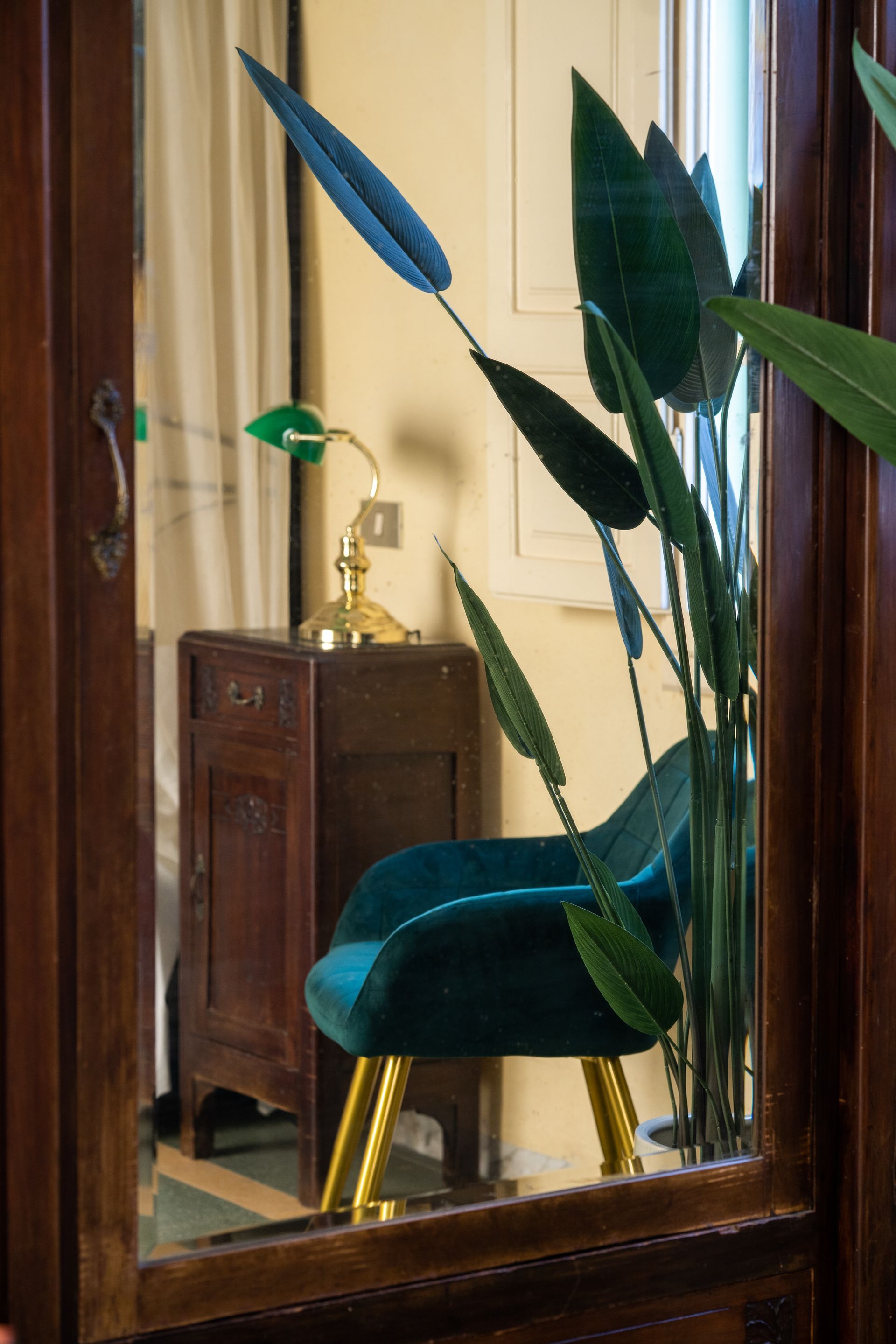 Green velvet chair, gold legs, behind a dark wooden frame; tall green plant, lamp on small table.
