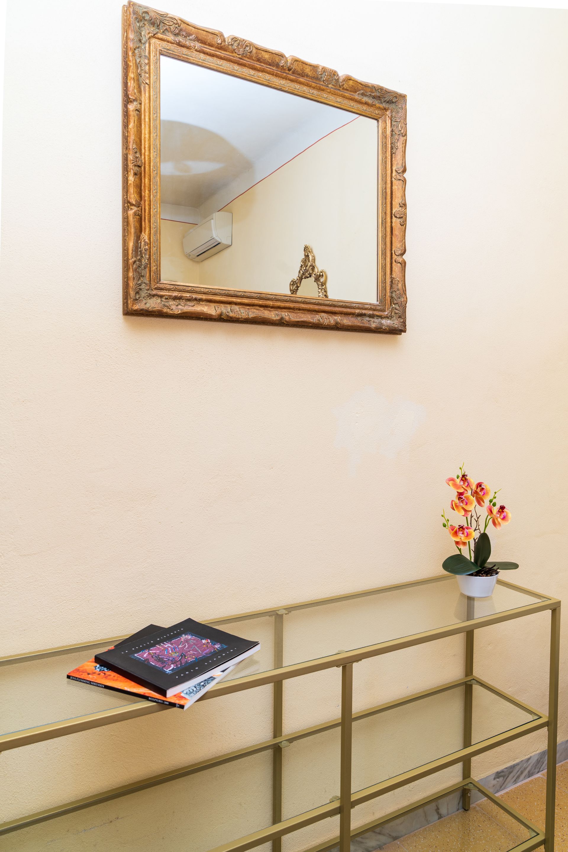 A hallway with a gold-framed mirror over a glass-top table with books and a flower.