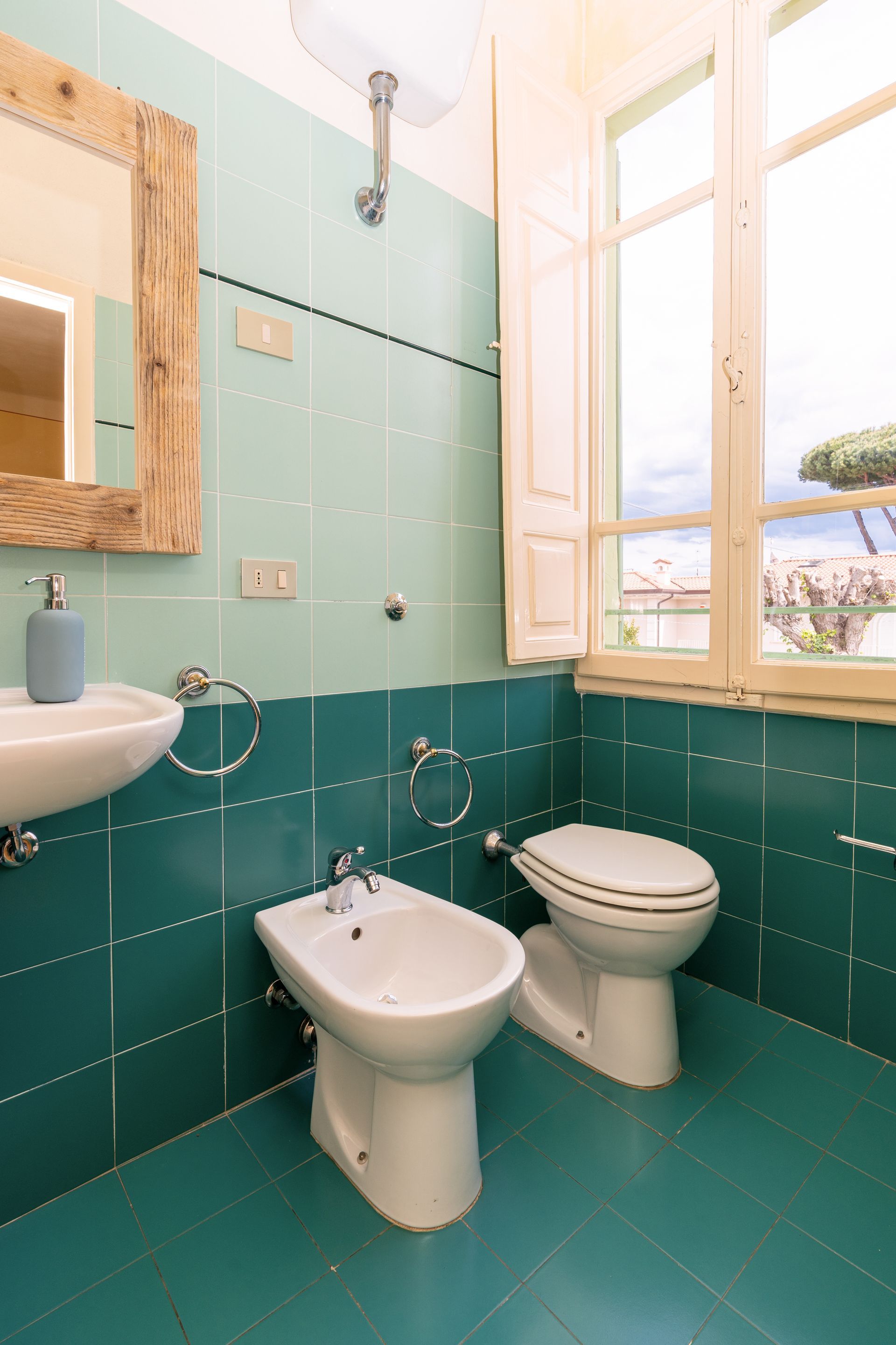 Bathroom with teal tile walls and floor, toilet, bidet, sink, mirror, and window.