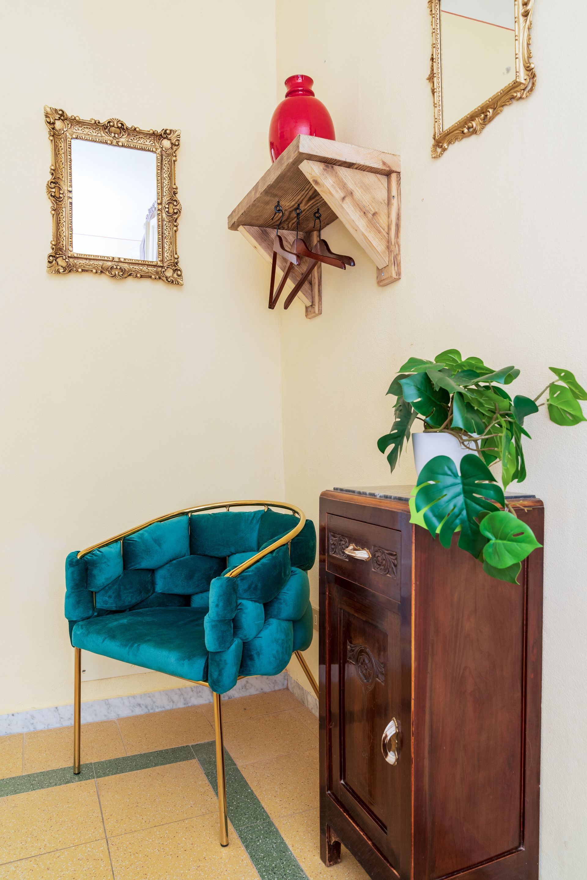 Teal armchair, wooden cabinet, and gold framed mirrors in a corner with a plant and decorative shelf.
