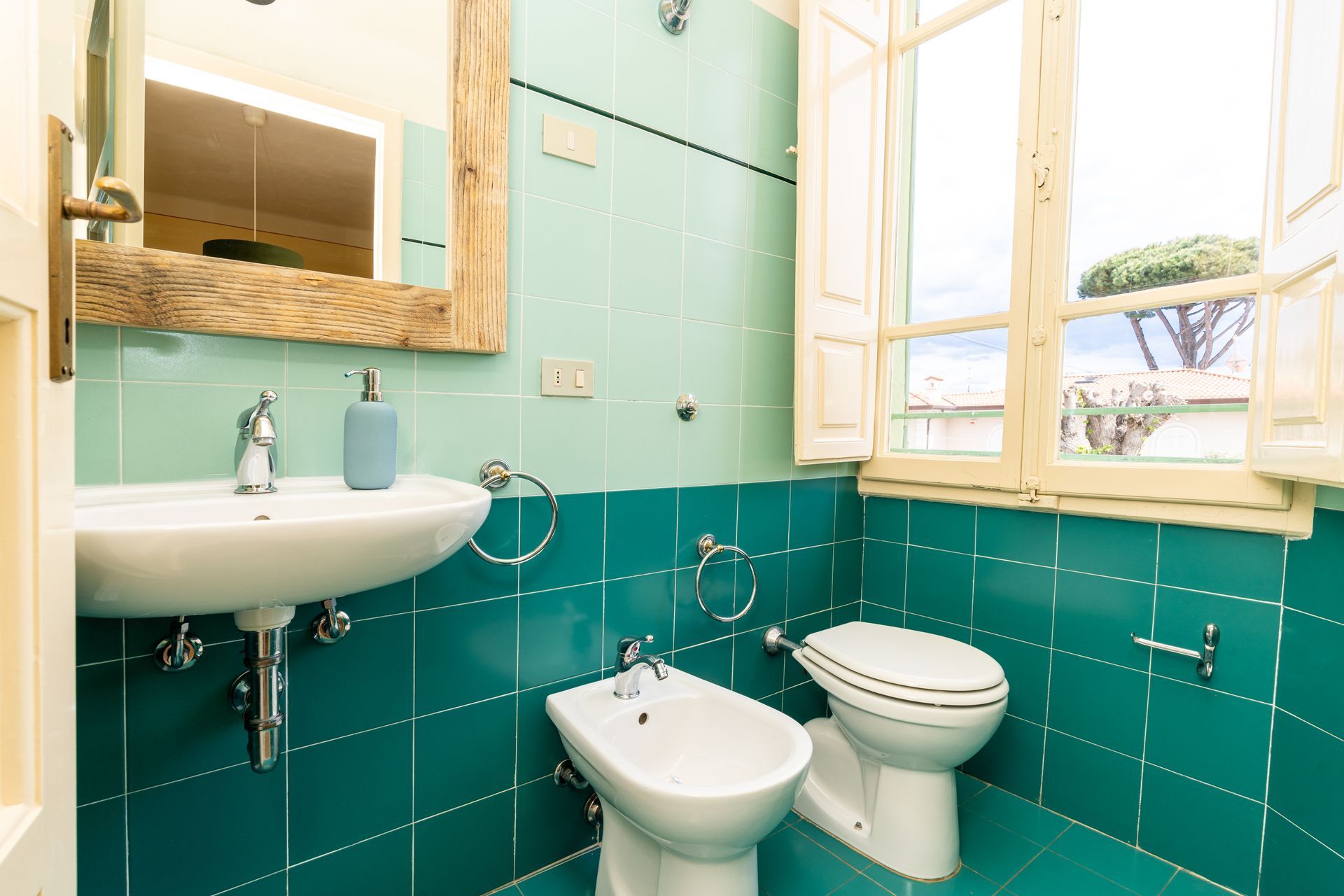 Bathroom with teal tiles, white sink, toilet, and bidet. Wooden-framed mirror and window with outdoor view.