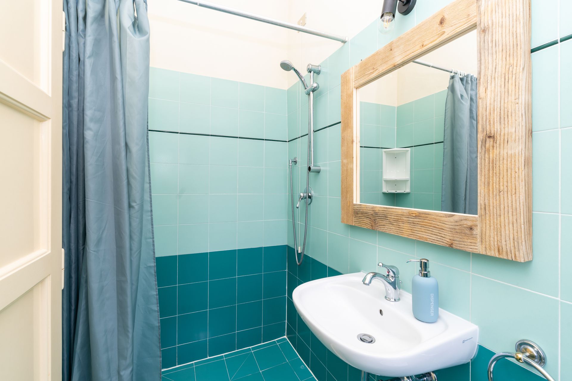Small turquoise bathroom with shower, sink, and wood-framed mirror.