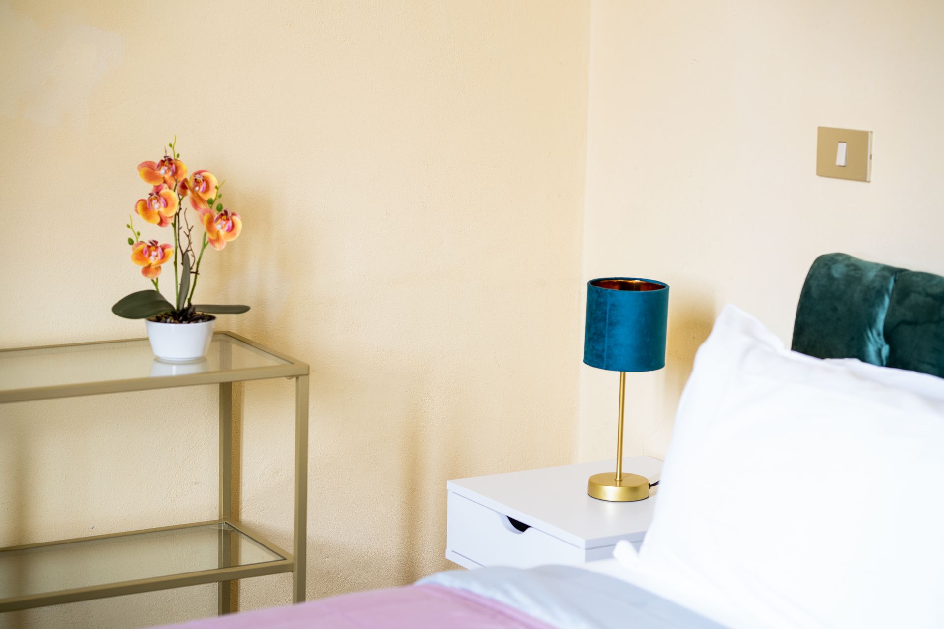 Bedroom corner with a bed, nightstand with lamp, and a plant on a glass table.