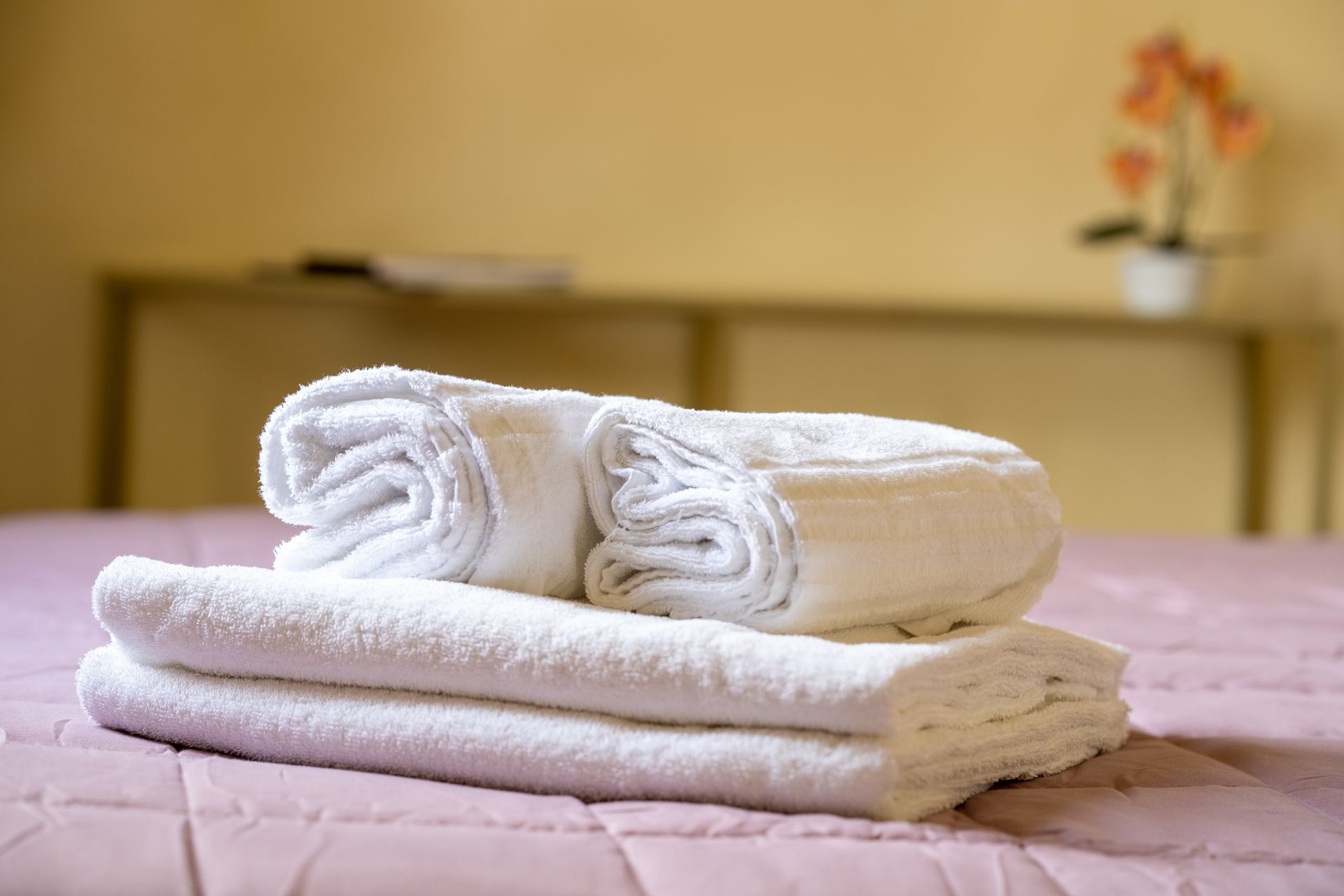 White rolled and folded towels on a pink bed, with a shelf and plant in the blurry background.