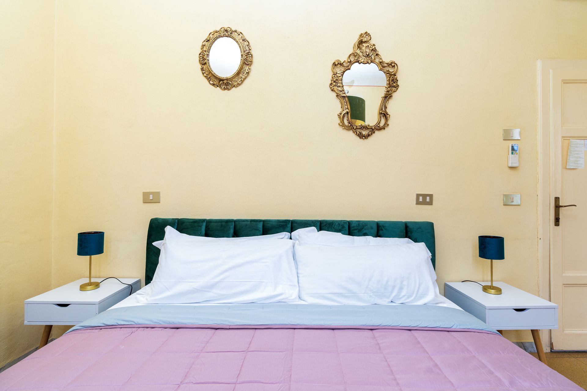 Bedroom with a bed, nightstands, lamps, mirrors, and door. The bed has a green headboard and pink blanket.