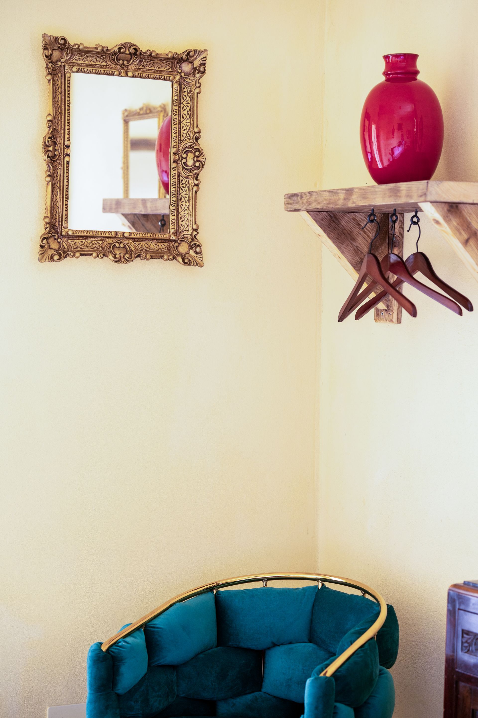A gold-framed mirror and a vibrant red vase on a wooden shelf in a room with a teal armchair.