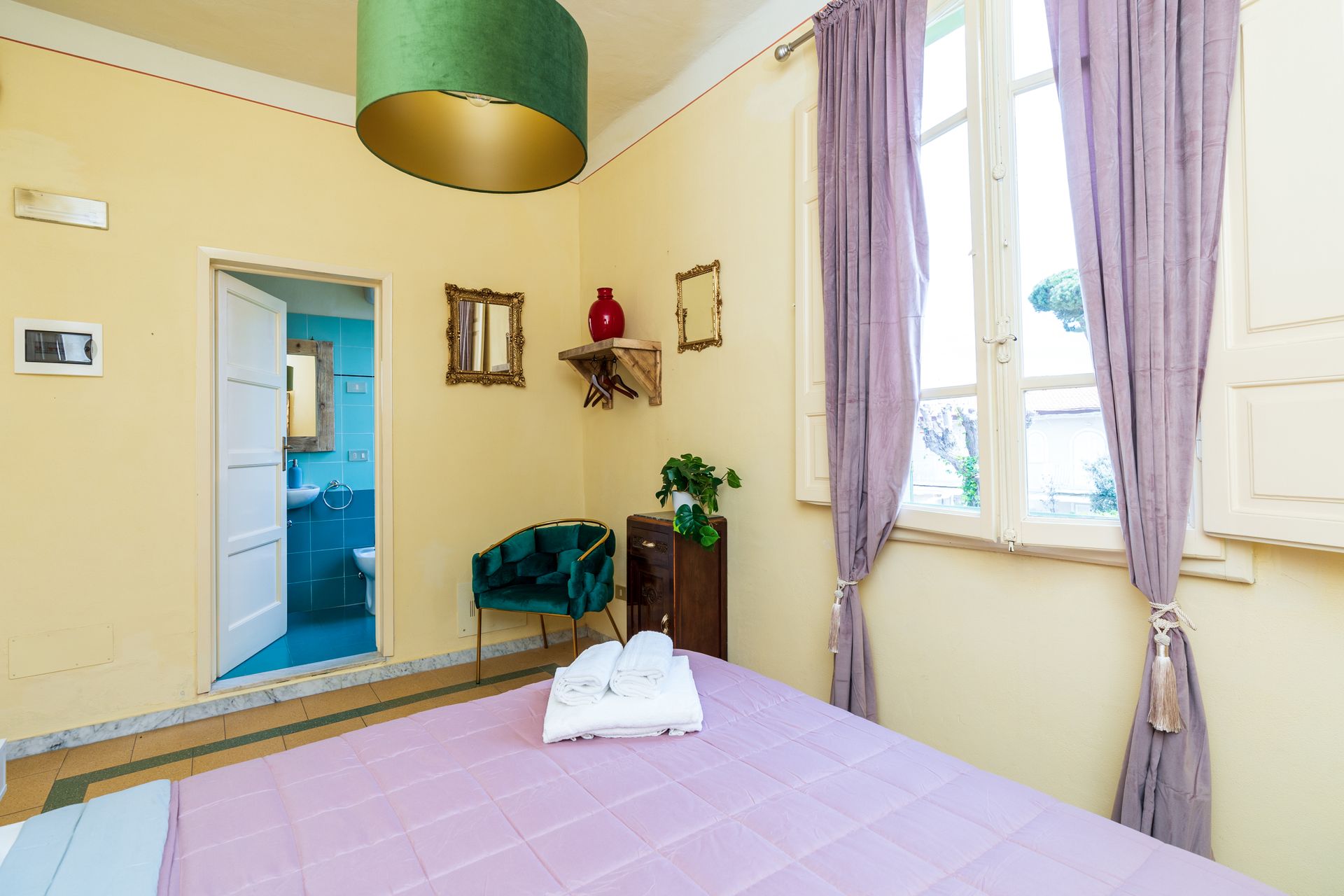 Bedroom with lilac bedspread, purple curtains, and a door to a blue bathroom.