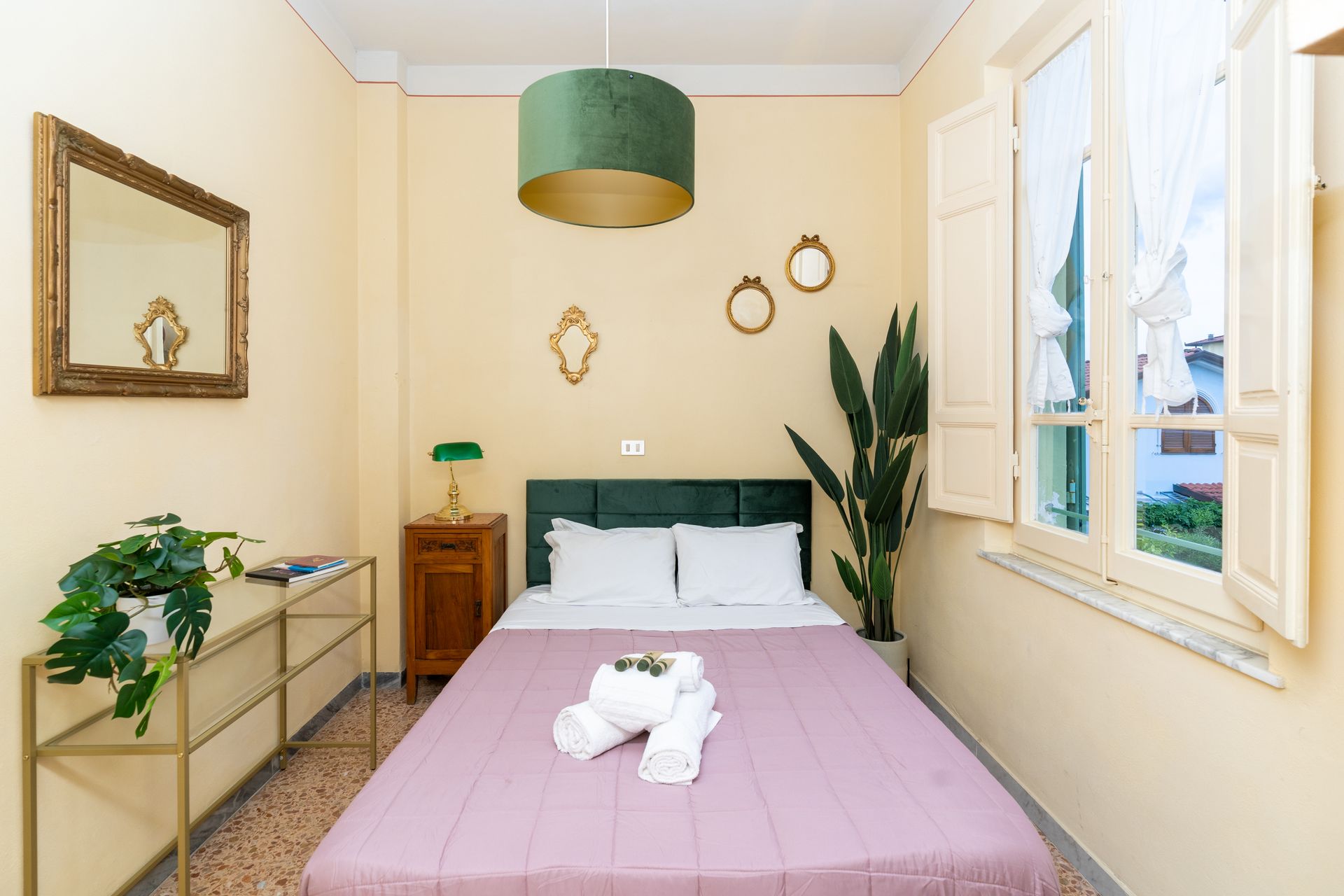 Bedroom with a bed, nightstand, mirror, and window with shutters. Green pendant lamp and plant.