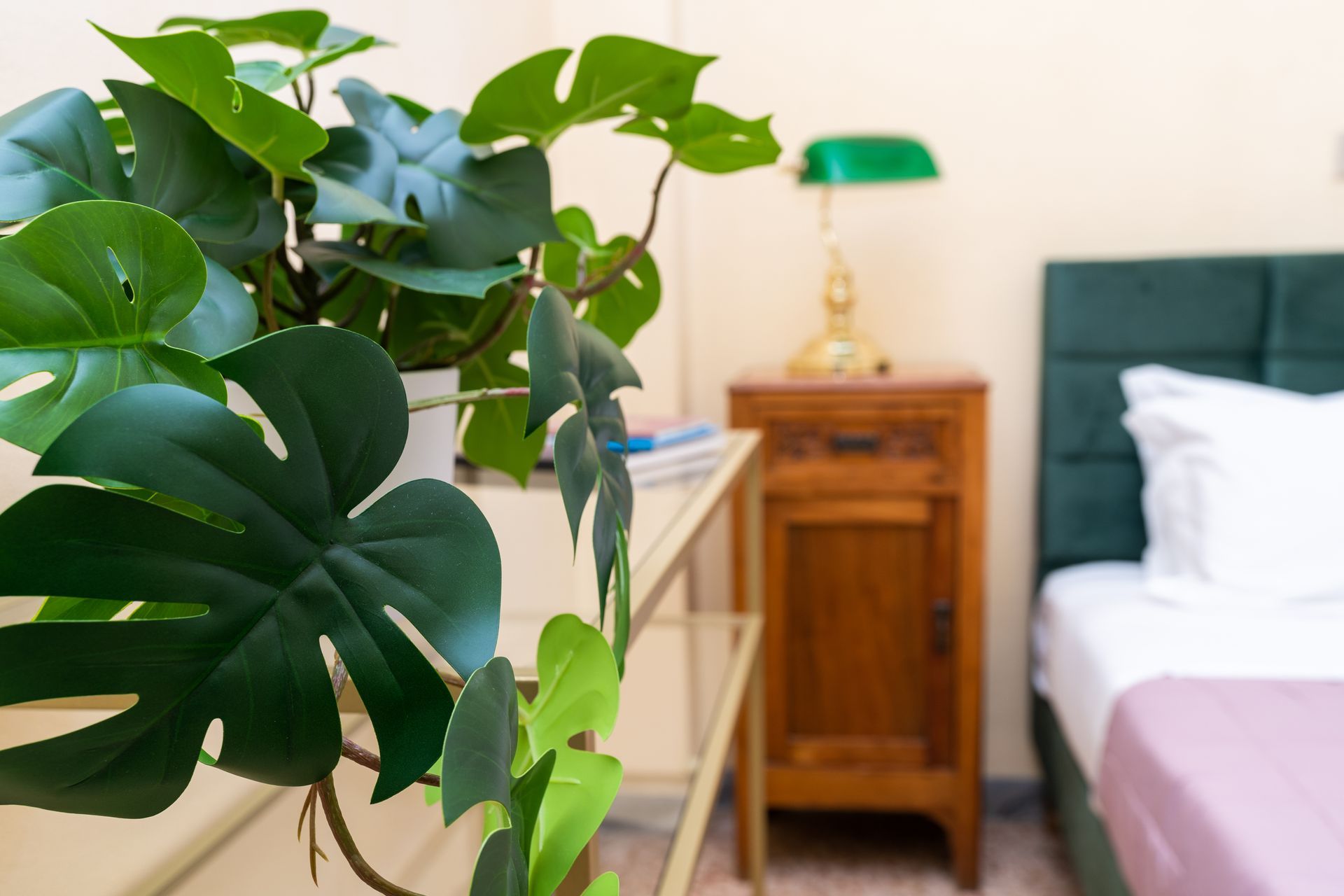 A bedroom scene: Monstera plant in focus, nightstand with green lamp, bed with pink sheet.