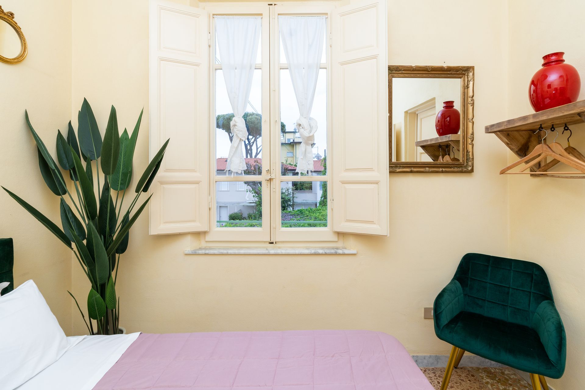 Bedroom with window, plant, mirror, chair, and red vase. Pale yellow walls and pink bedspread.