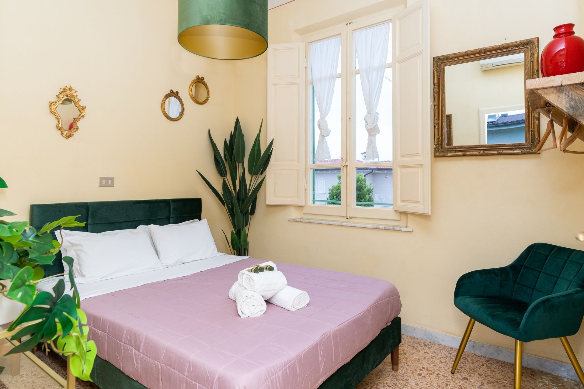 Bedroom with double bed, green velvet headboard and chair, window with shutters, decorative mirrors, cactus, and hanging light.