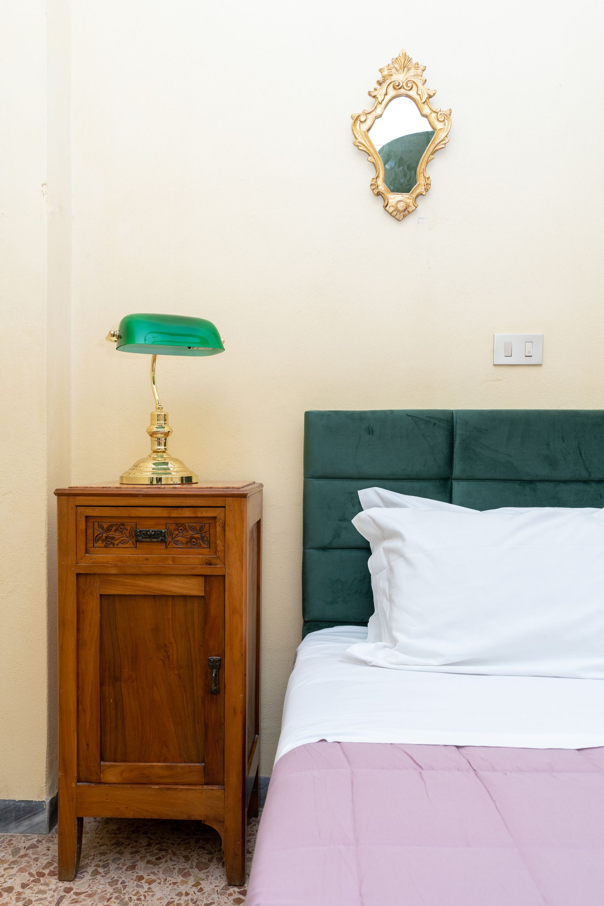 Bedside table with green lamp next to bed with green headboard and small gold mirror.