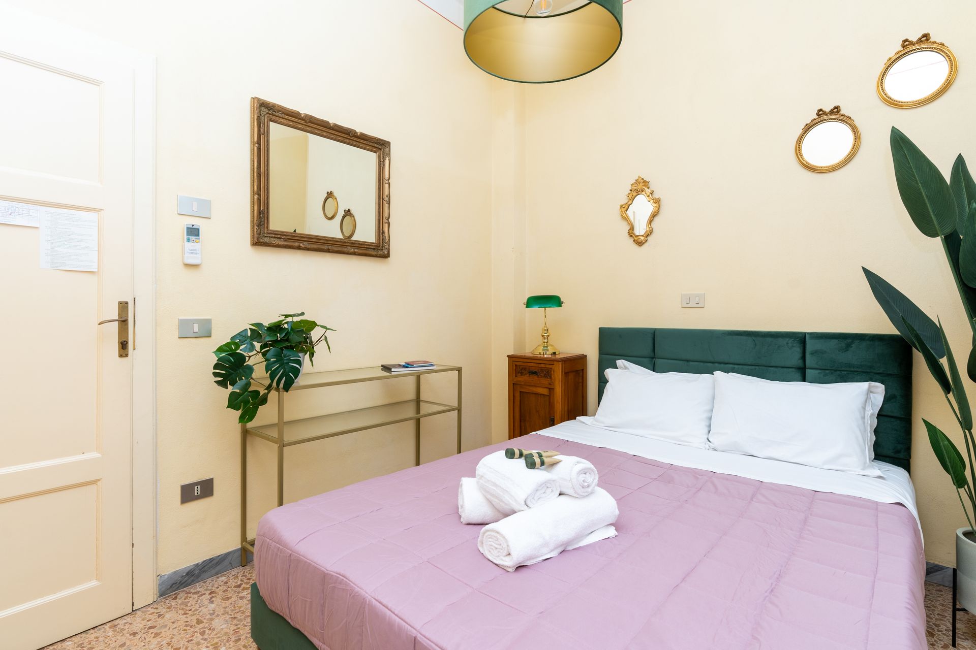 Bedroom with a bed, small side table, mirror, and decorative plants. Pale yellow walls, pink bedspread, and green headboard.