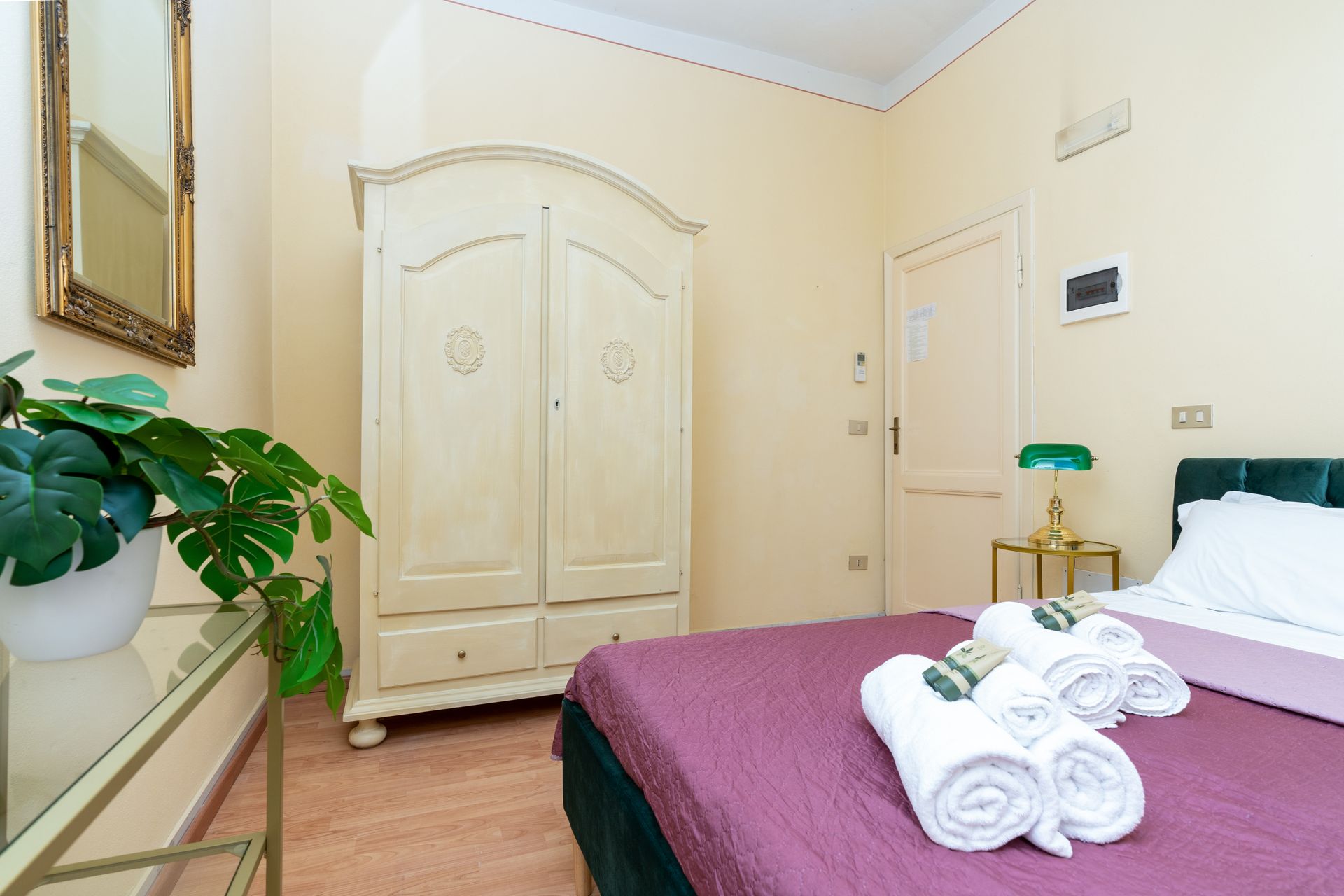 Bedroom with bed, wardrobe, mirror, and side table with lamp. Towels folded on bed.