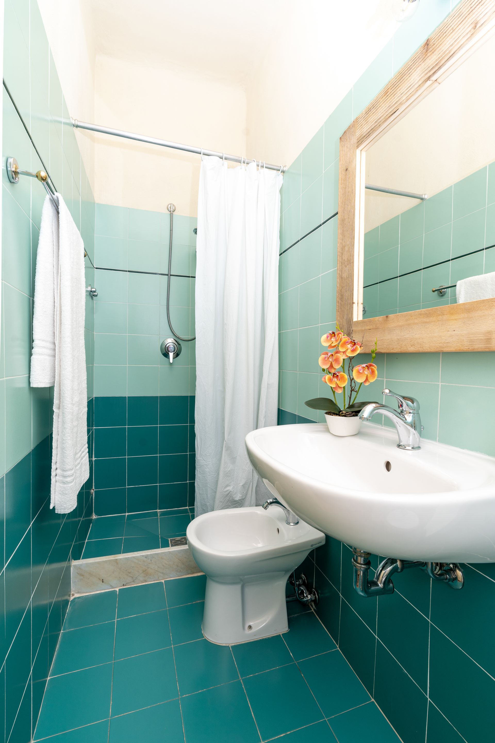 Bathroom with teal tile, white sink and bidet, shower curtain, and a mirror with a wooden frame.