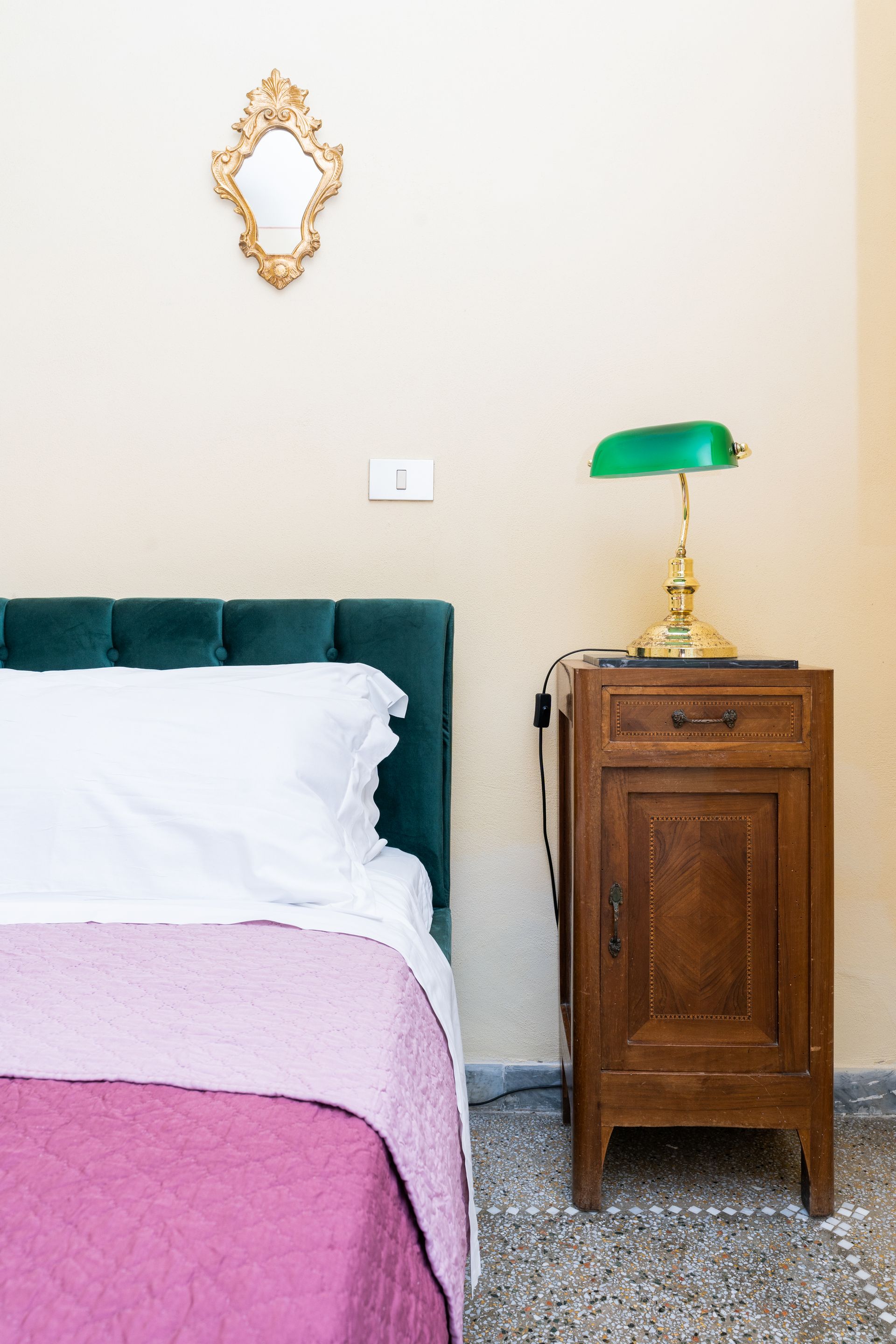 Bedroom with green headboard, bed with pink cover, bedside table, and ornate mirror.