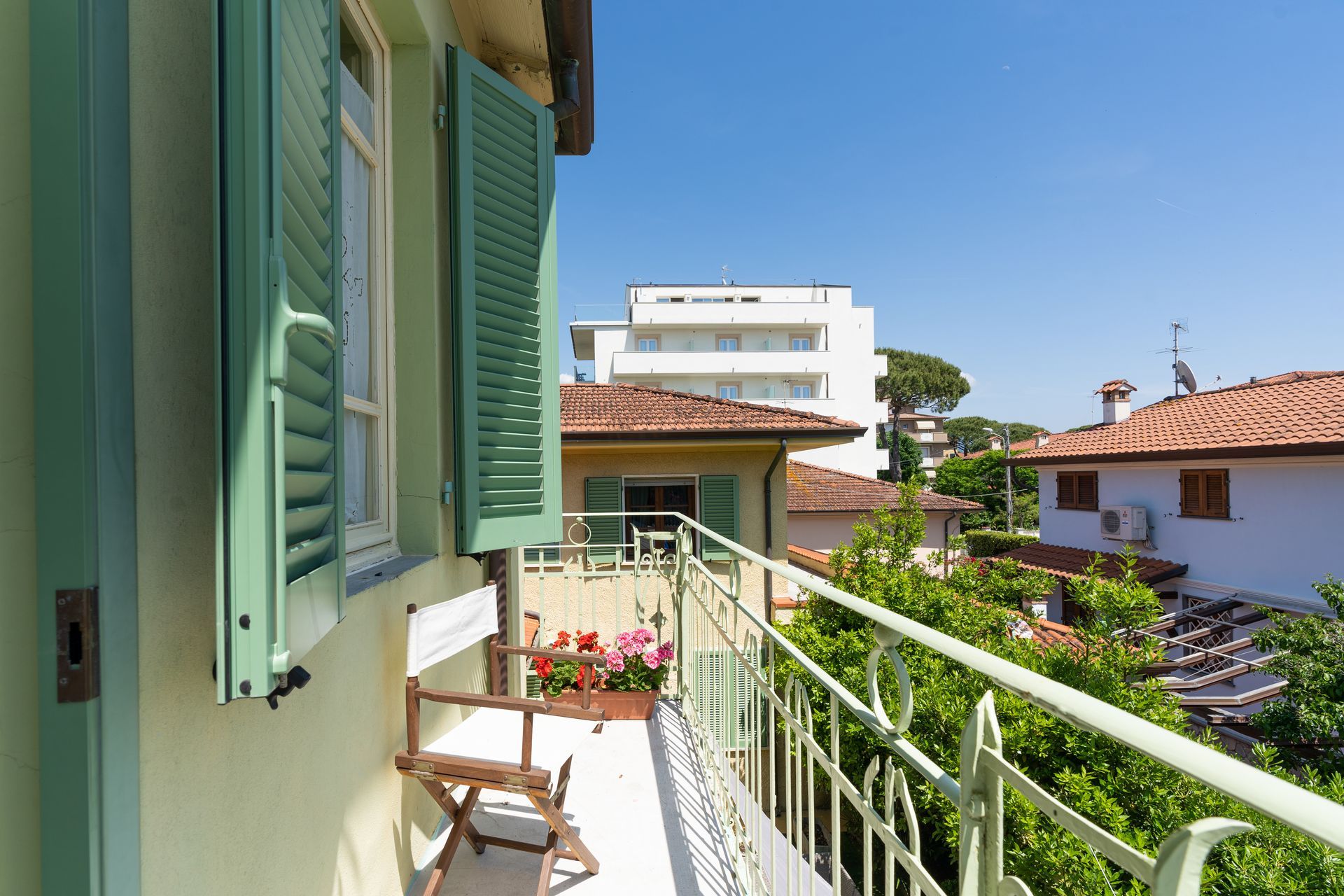 Balcony with green shutters, a small white bench, and a view of buildings and trees under a clear blue sky.