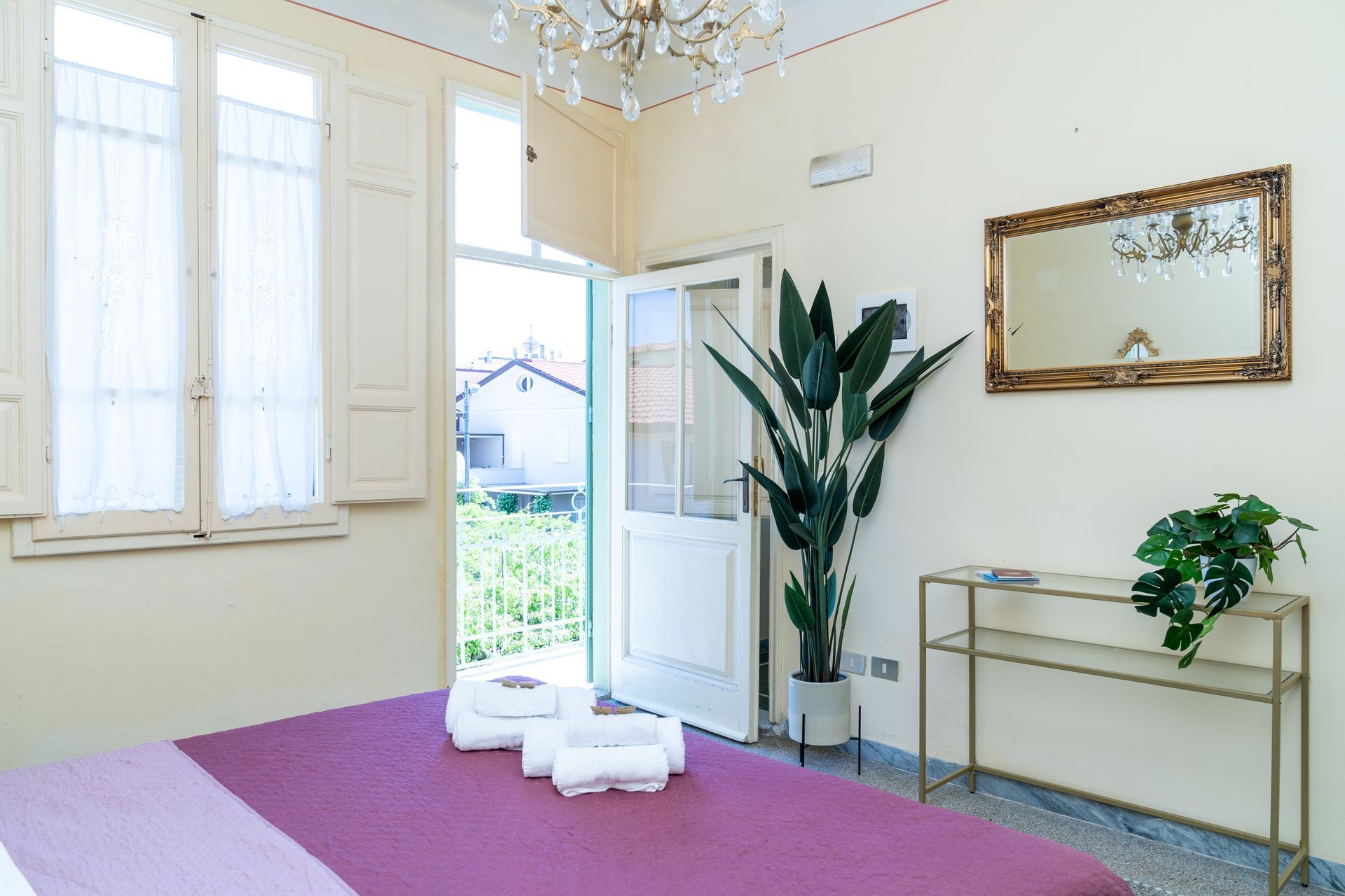 Bedroom with open door to a garden. Bed has purple cover, white towels, and a chandelier.