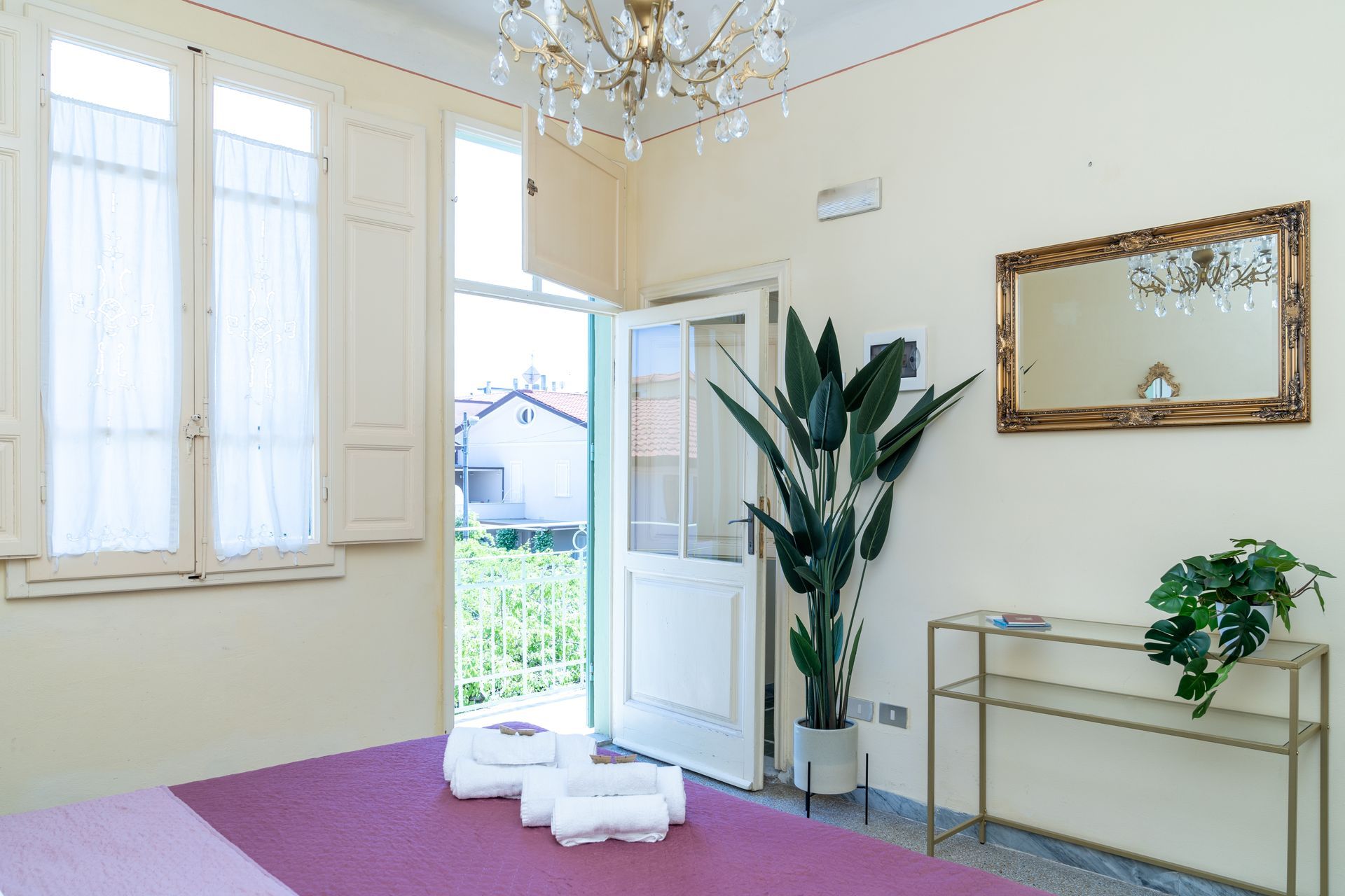 Bedroom with balcony, bed, open door, window, mirror, chandelier, and potted plants.