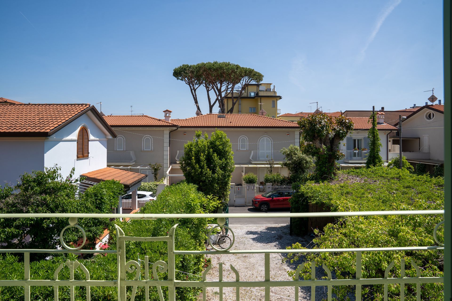 View of houses with red roofs and a car on a cobblestone driveway, framed by a green wrought iron balcony and lush foliage under a clear blue sky.