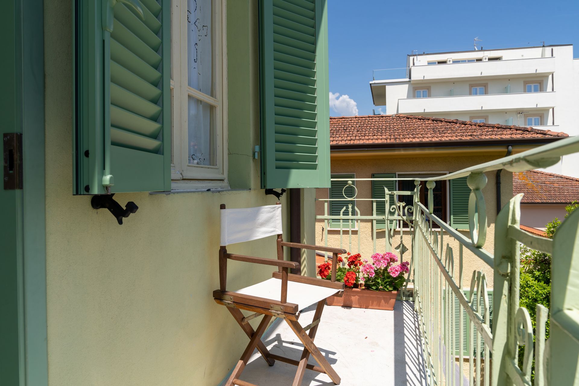 Balcony with folding chair, flowers, and green shutters; buildings in the background.