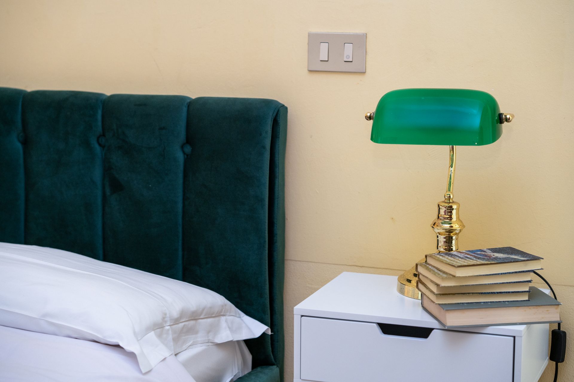 Emerald green velvet headboard, white pillow, bedside table with green lamp and books.