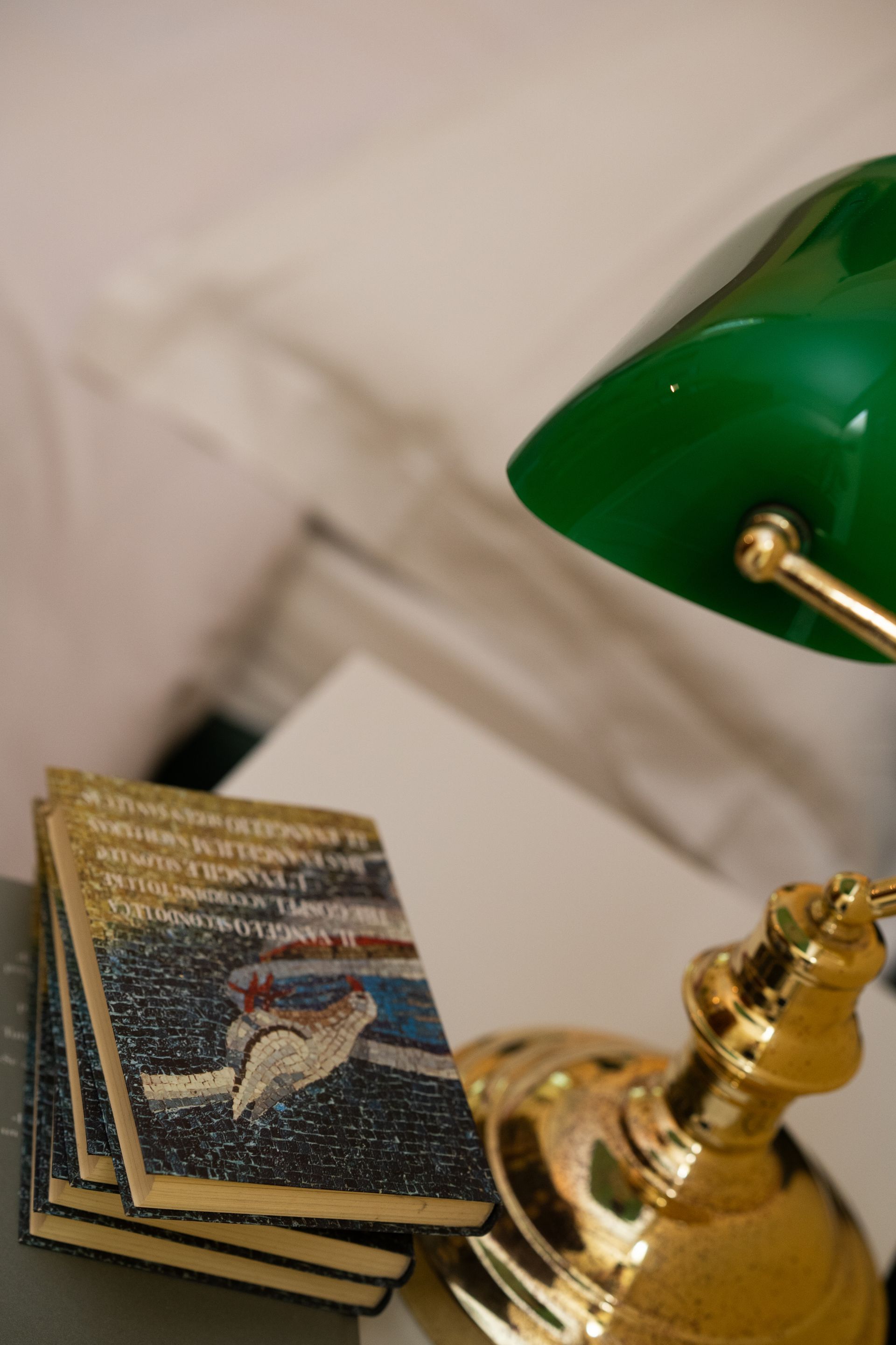 Stack of books with a bird illustration, under a green-shaded brass lamp on a table.