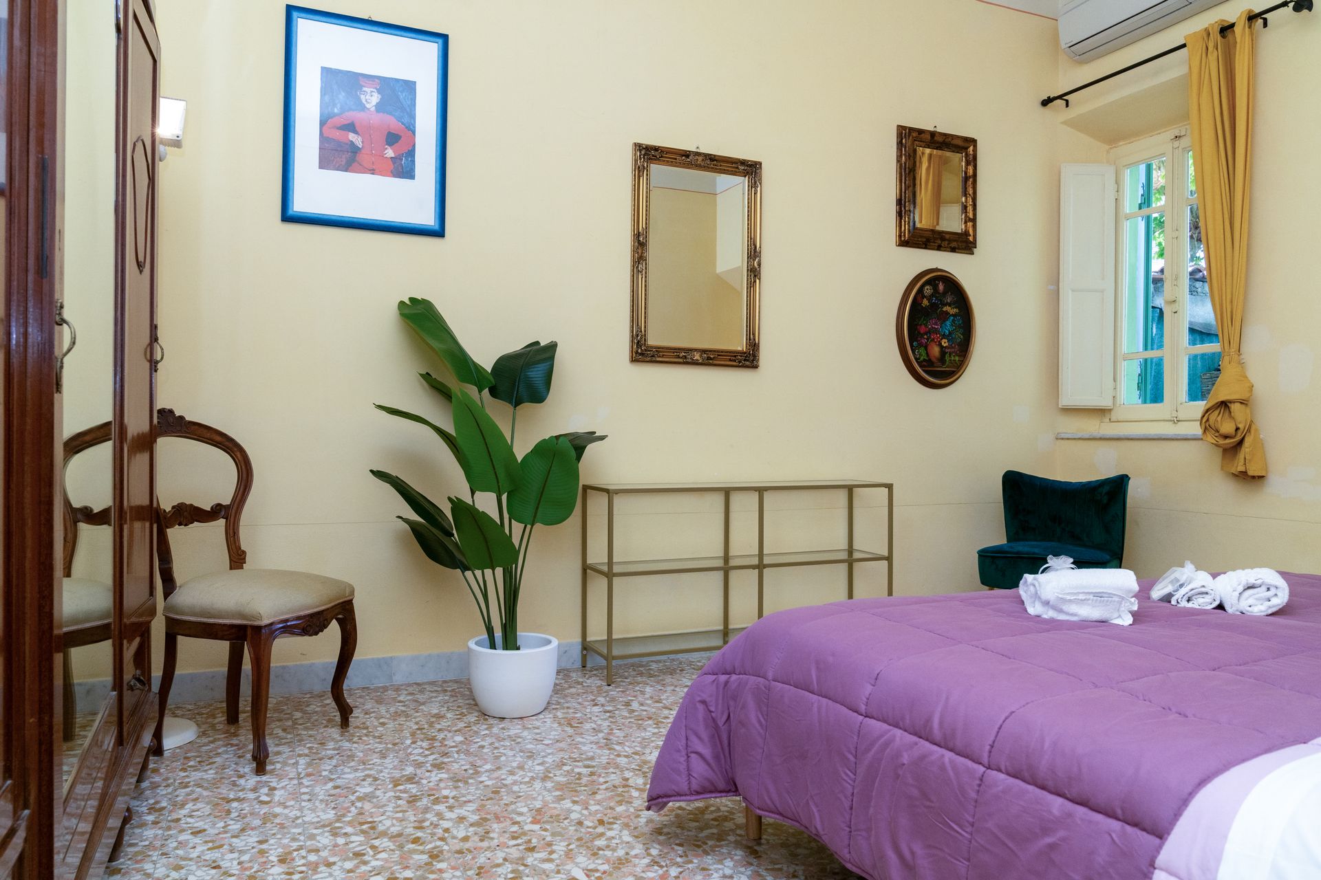 Bedroom with bed covered in purple, framed art, ornate mirror, and potted plant.