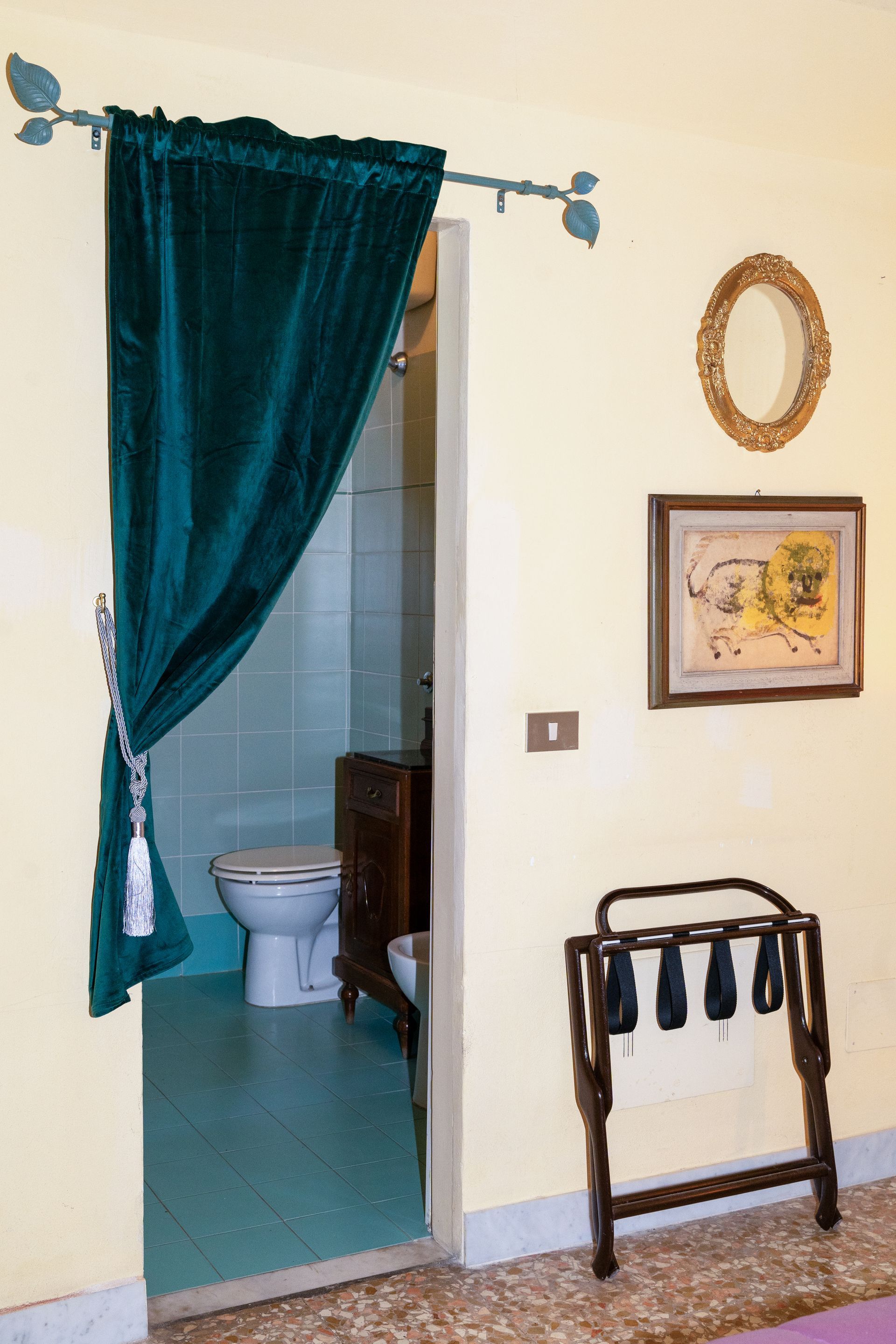 Bathroom doorway with teal curtain, luggage rack, and framed art on a yellow wall.