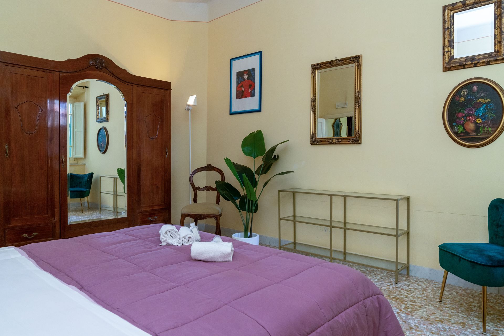 Bedroom with large wooden wardrobe, bed with purple cover, and gold-framed mirrors and art on the walls.