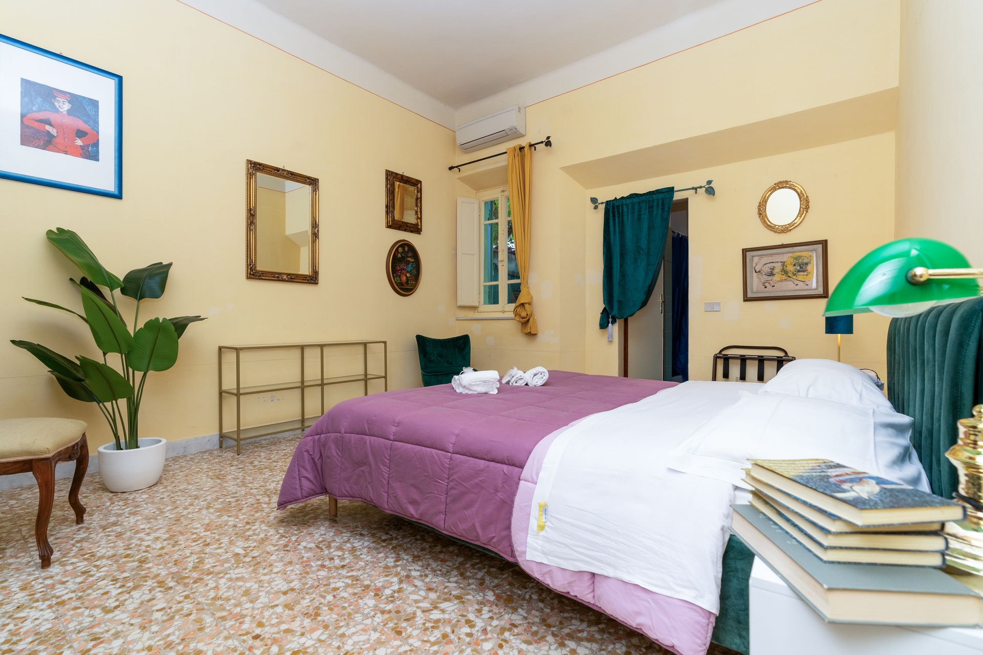 Bedroom with bed, books, green lamp, art, and plant. Yellow walls, patterned floor.