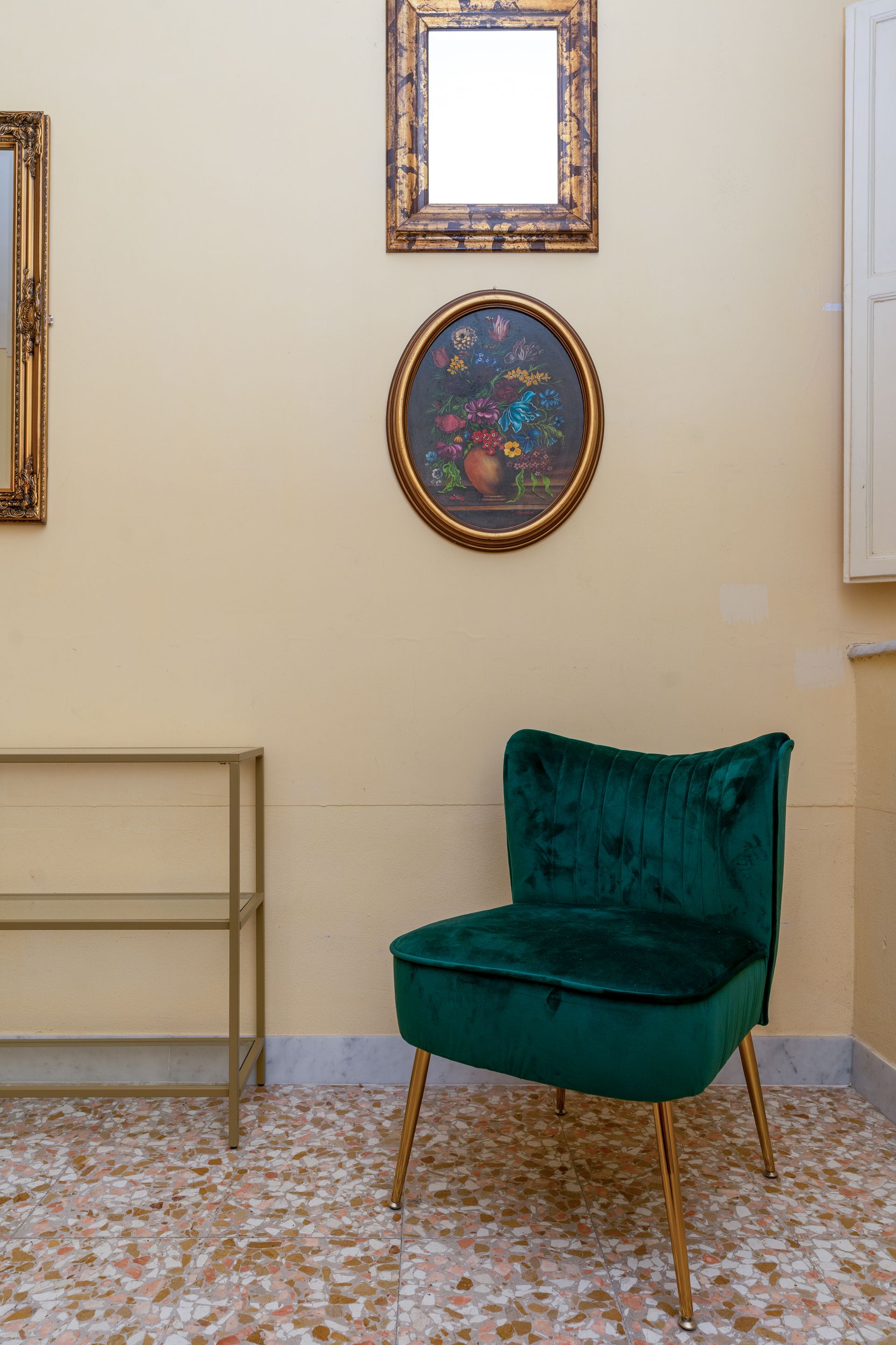 Green velvet chair with gold legs, next to a gold shelving unit. Artwork hangs on a cream-colored wall.