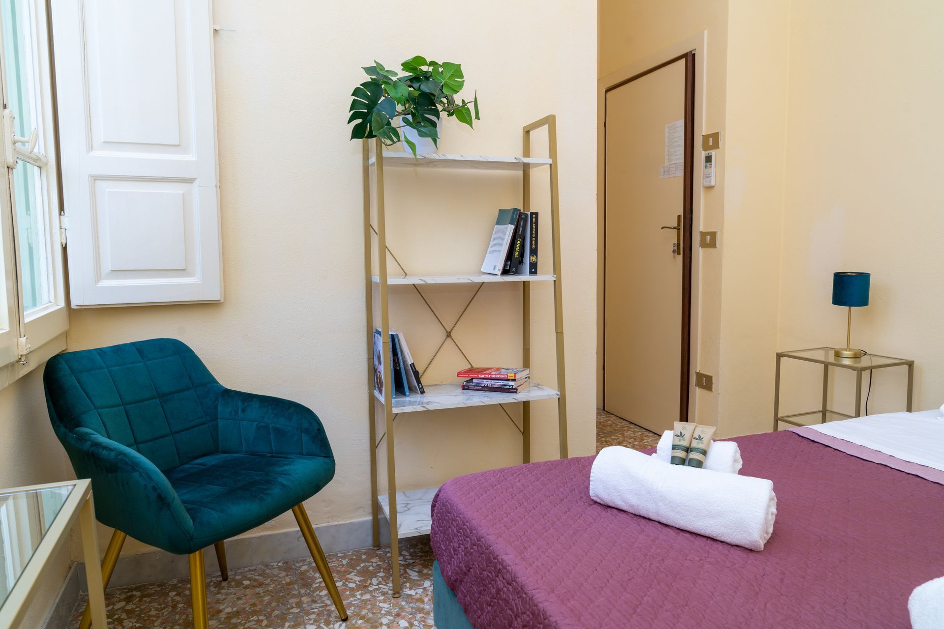 Bedroom with teal chair, bookshelf, and bed with white towels. Light walls and door.