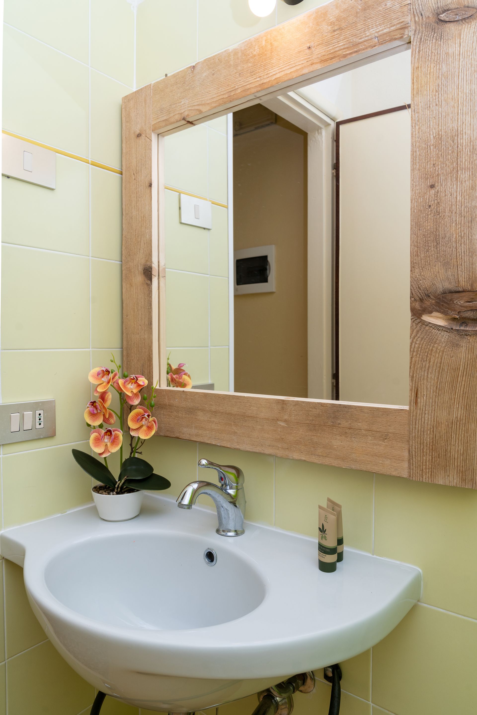 Bathroom sink with wooden framed mirror, flower, and green tiled walls.