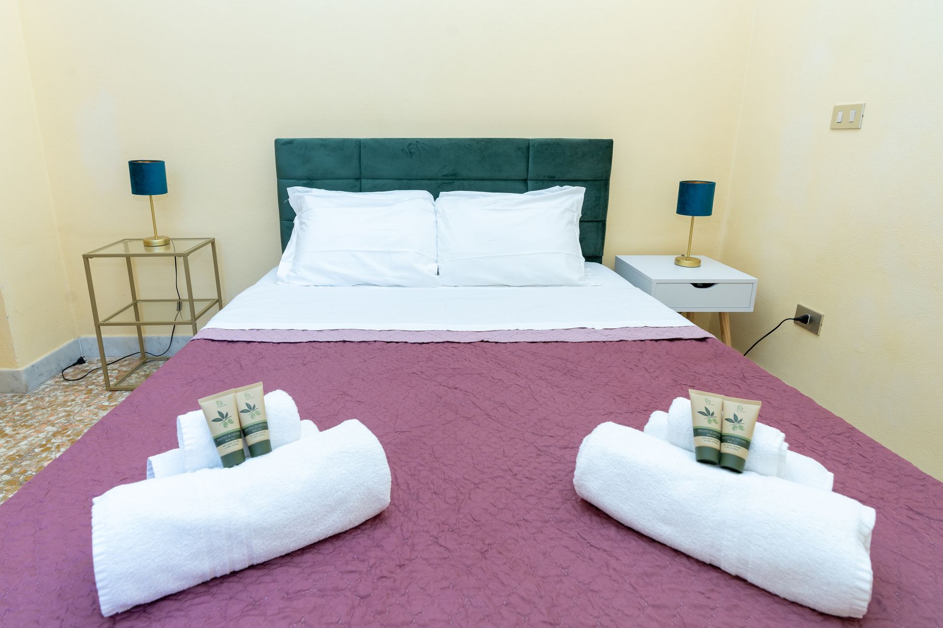 Bedroom with a double bed, purple cover, white sheets, teal lamps, and two rolled towels.