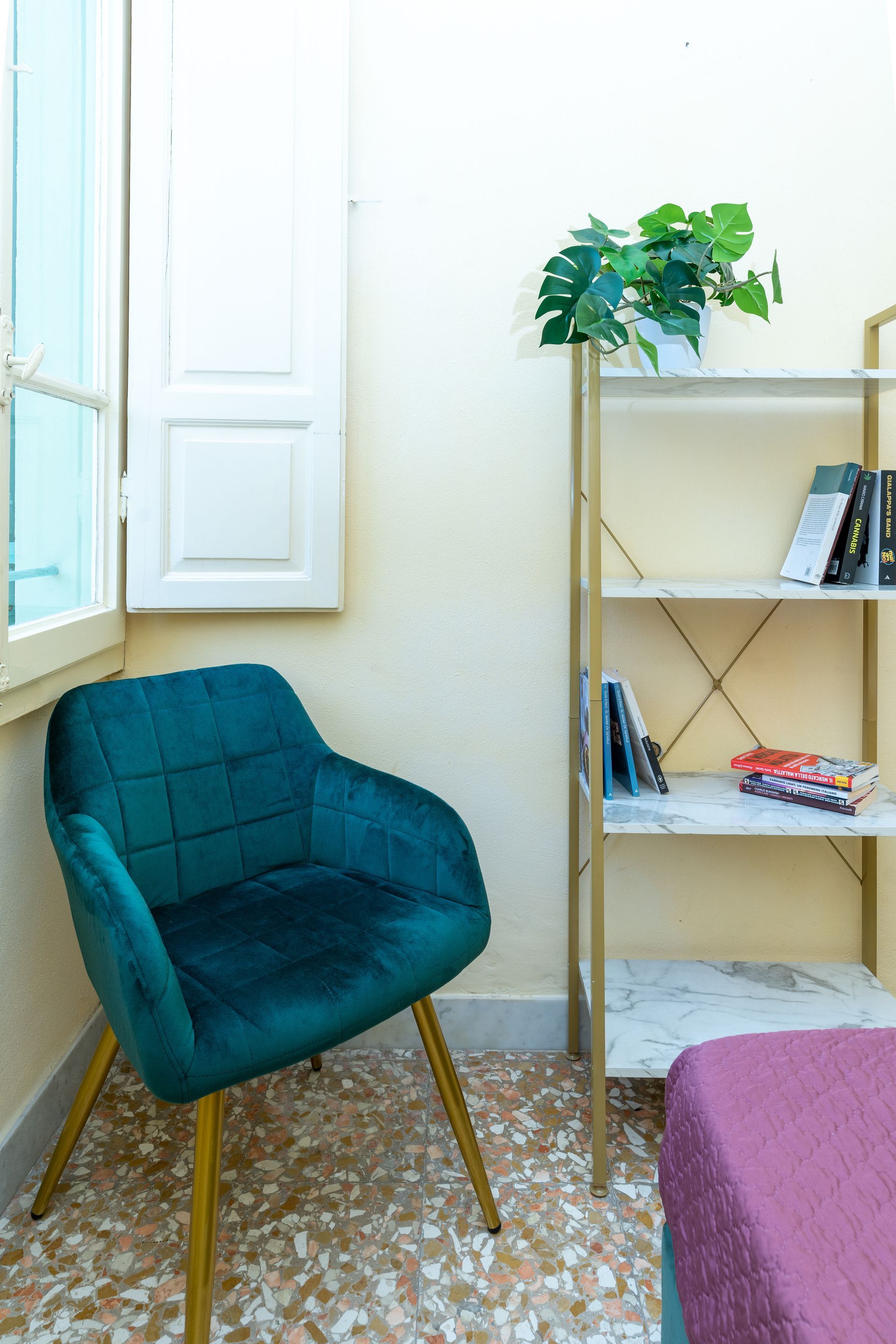 Teal velvet chair beside a bookshelf, next to a window with white shutters, in a corner.
