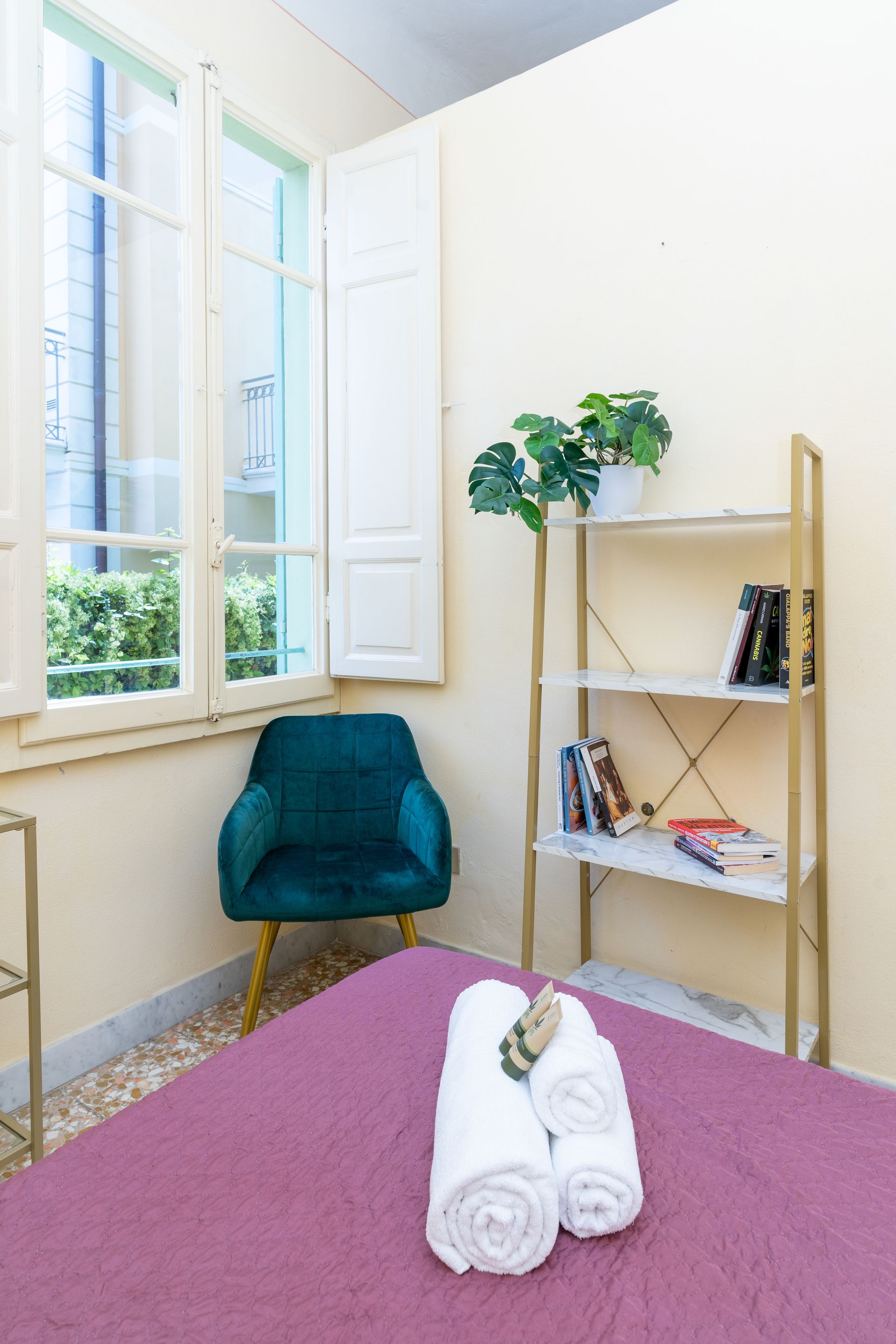 Bedroom with a bed, teal chair, window, shelves, and rolled white towels.