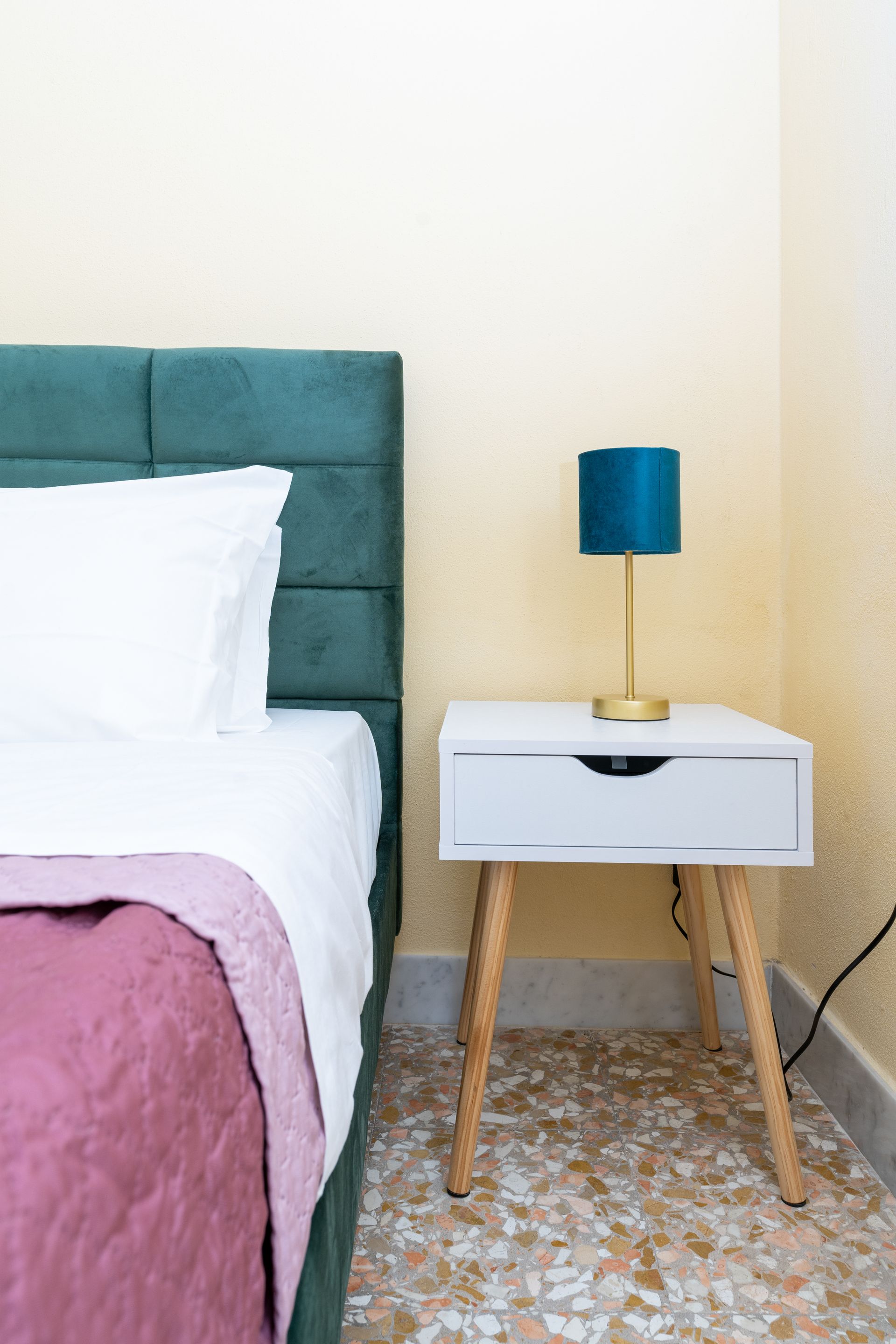 Bed with teal headboard, white bedding, pink quilt, white nightstand with lamp on patterned floor.