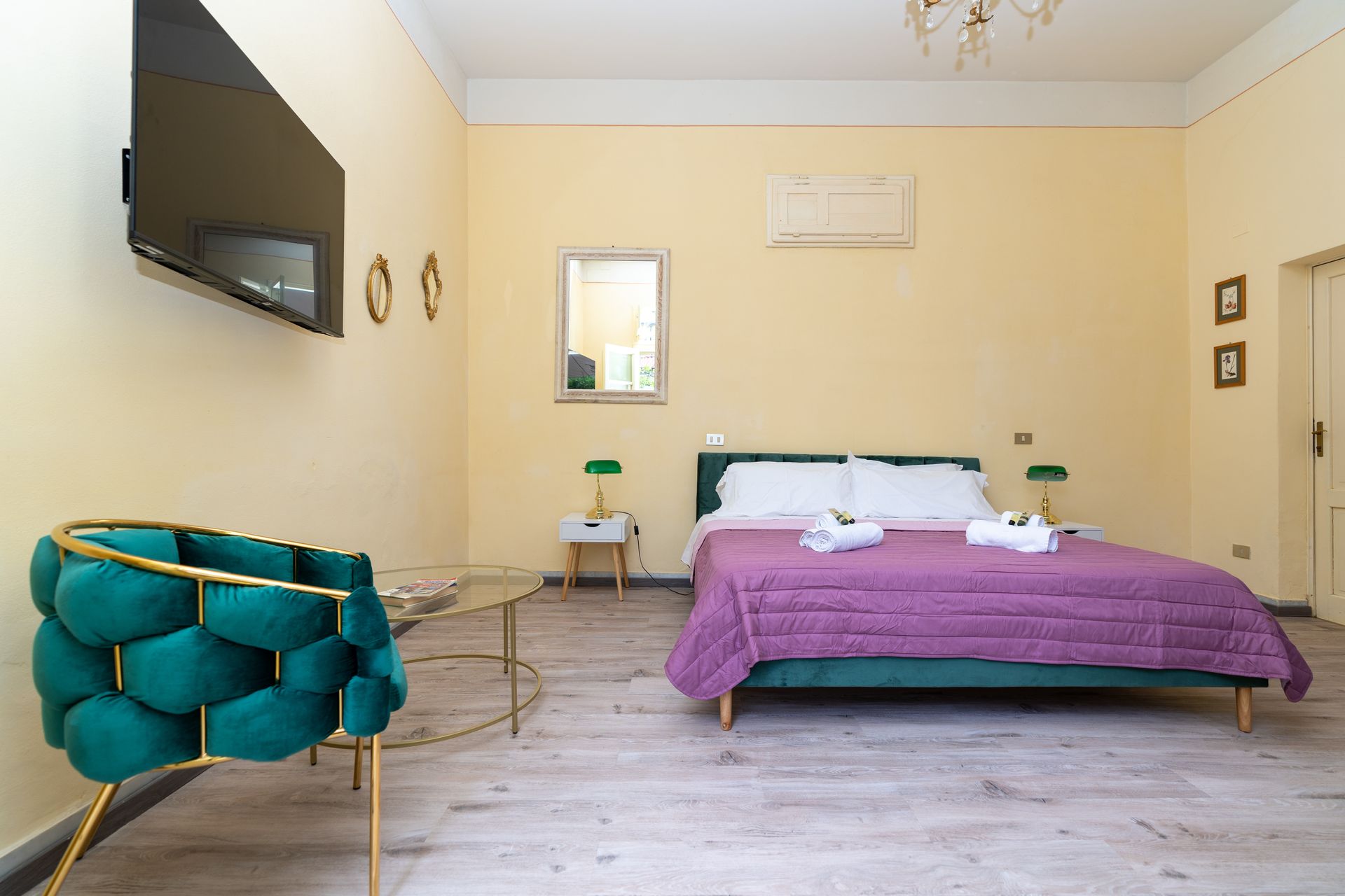 Bedroom with bed, purple cover, teal chair, and mounted TV. Pale yellow walls and light wood floors.