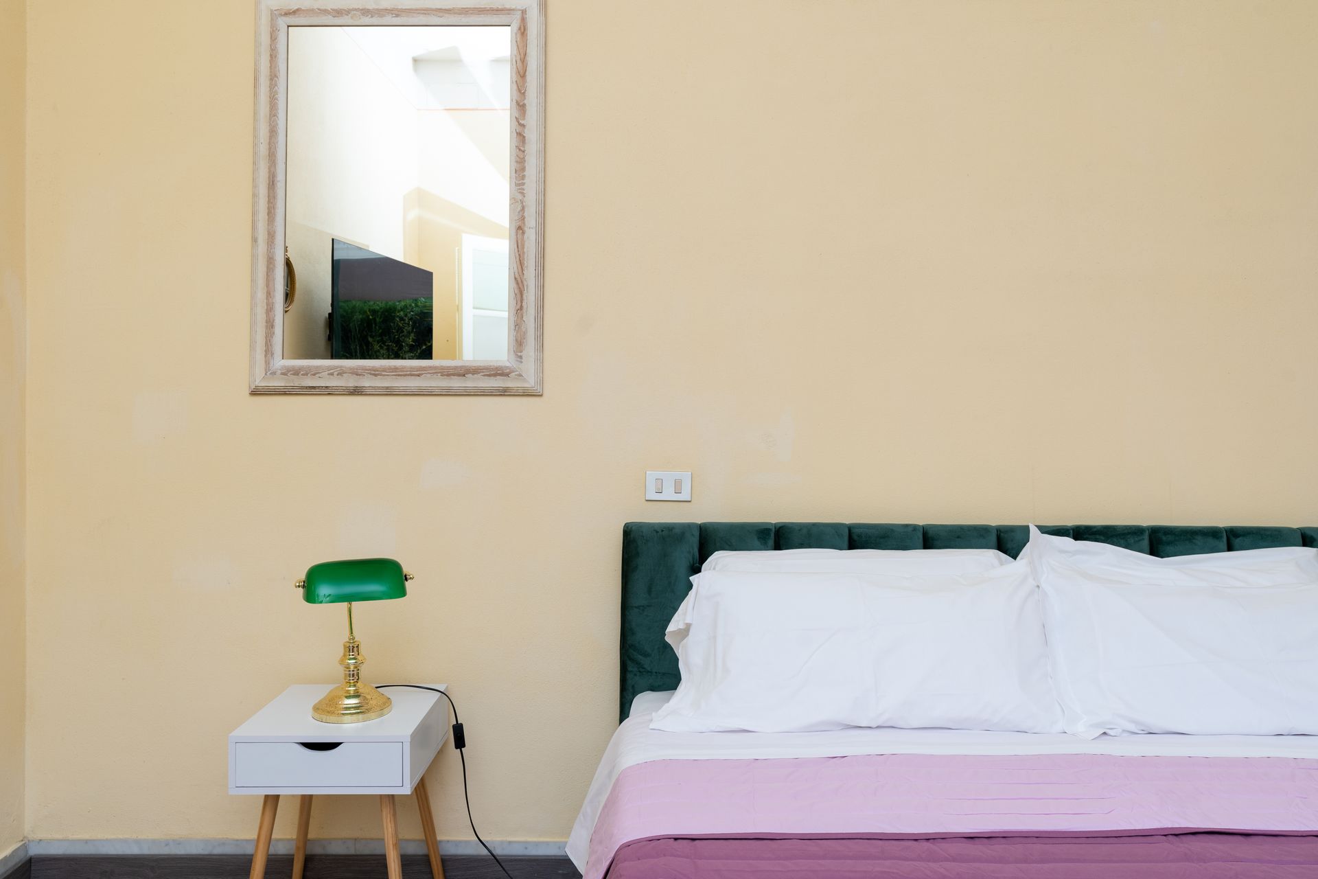 Bedroom with bed, nightstand with green lamp, and a mirror on a yellow wall.